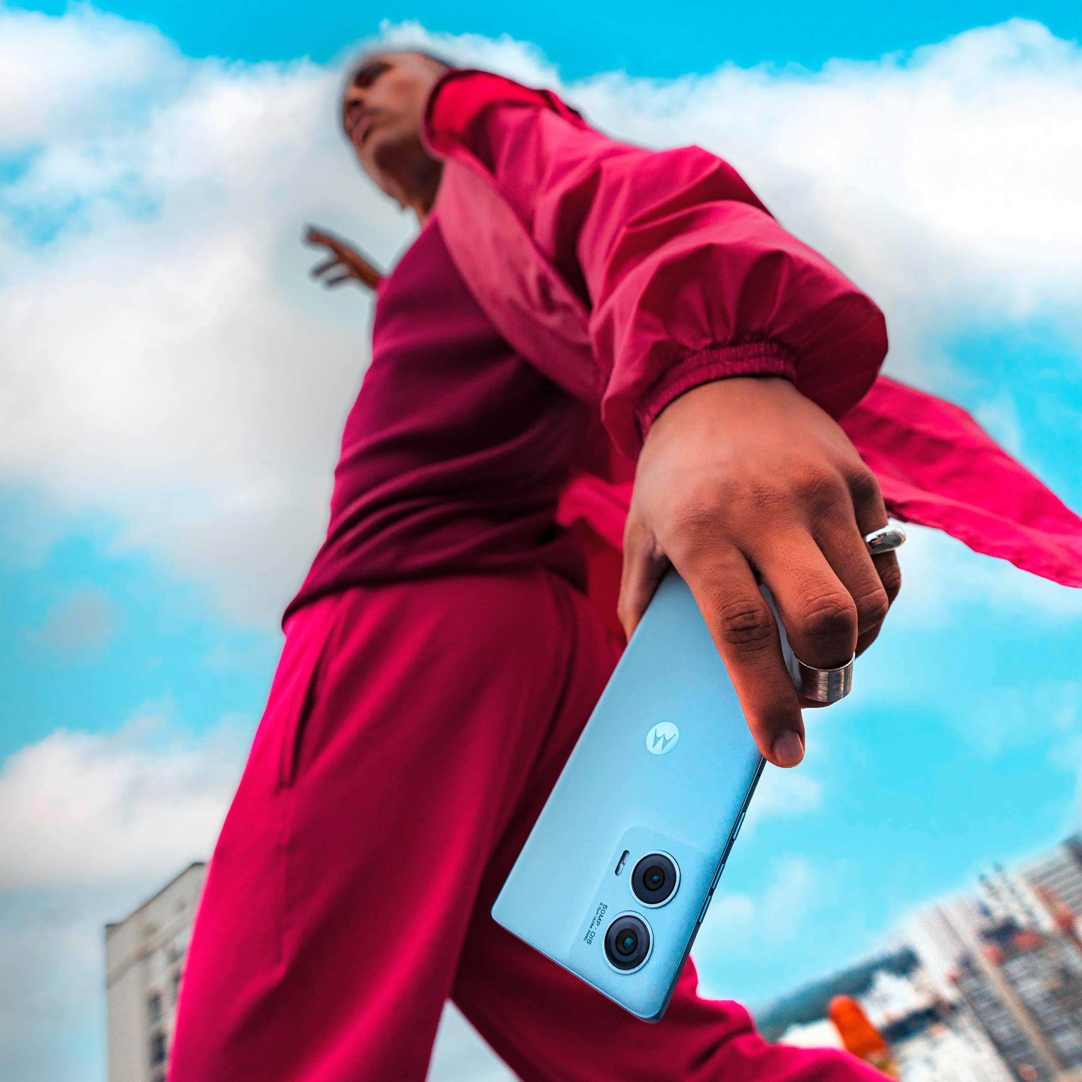 Osoba trzymająca jasnoniebieski smartfon z dwoma aparatami, na tle błękitnego nieba.