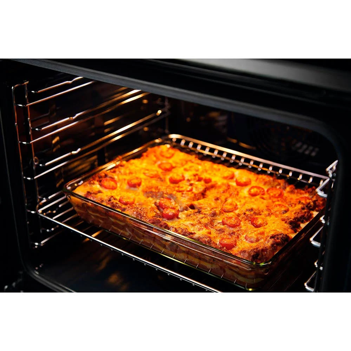 Szklane naczynie do pieczenia z lasagne w piekarniku, na ruszcie. Wnętrze piekarnika jest ciemne.