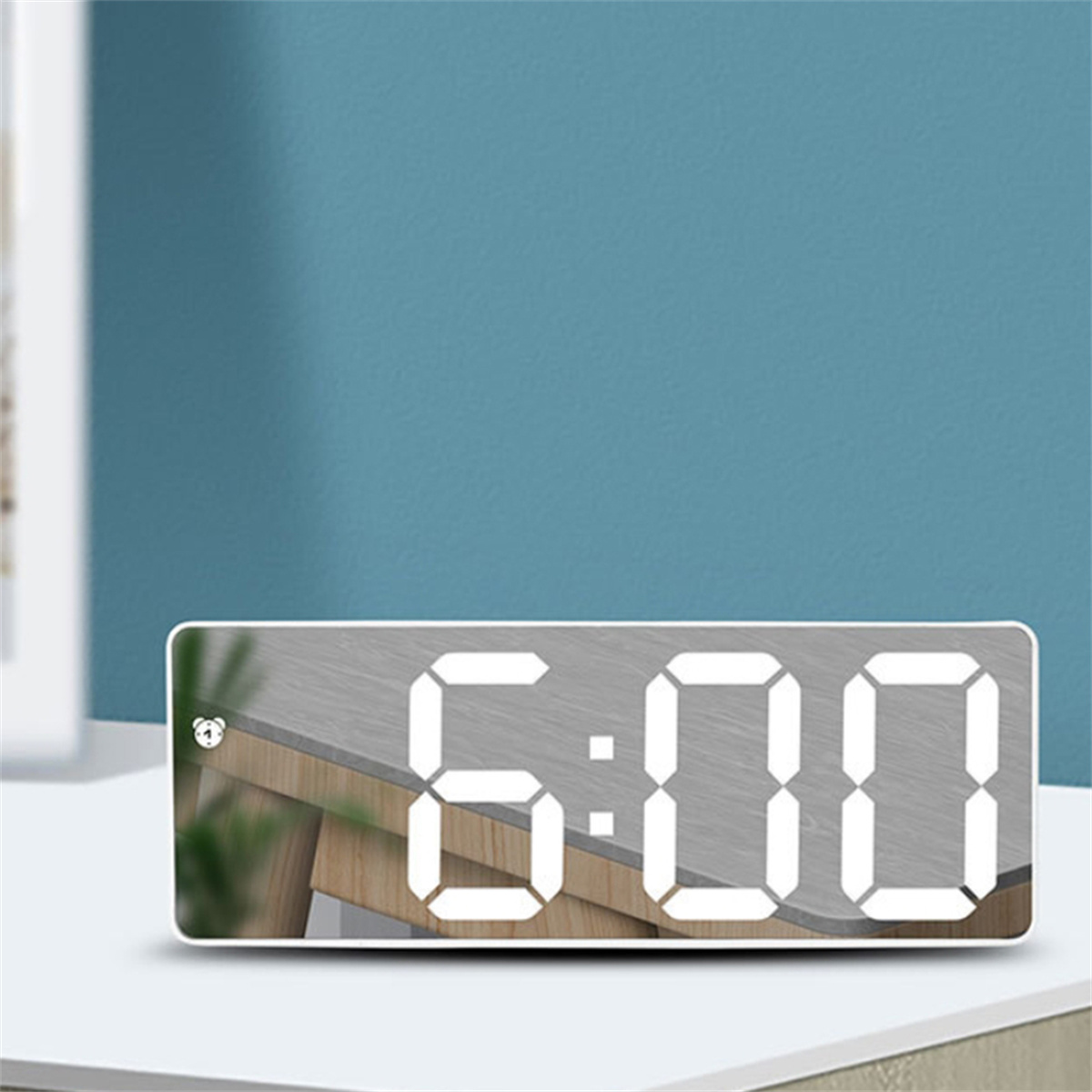 Digitaler Wecker mit der Anzeige 6:00 auf weißer Oberfläche, blauer Wandhintergrund.