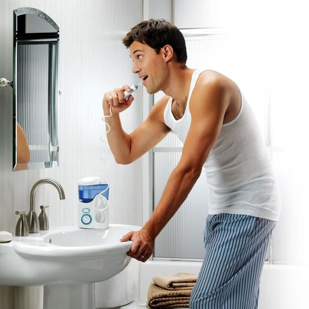Un hombre cepillándose los dientes sobre un lavabo en un baño. Un irrigador bucal está en el lavabo.
