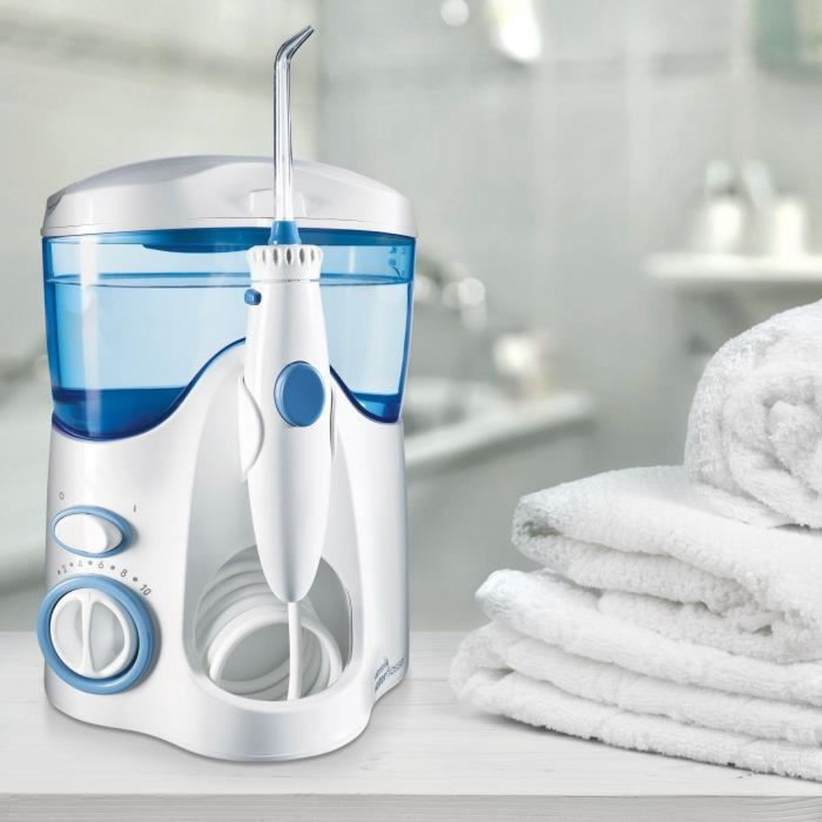 Irrigador bucal blanco y azul, depósito de agua y cabezal adjunto, con toallas blancas en el fondo.