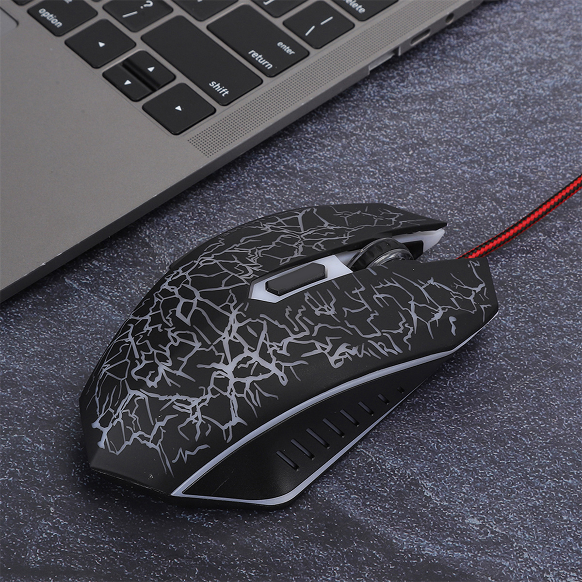 Eine schwarze Gaming-Maus mit weißem Rissdesign und einem roten Kabel. Sie liegt auf einer dunklen strukturierten Oberfläche.