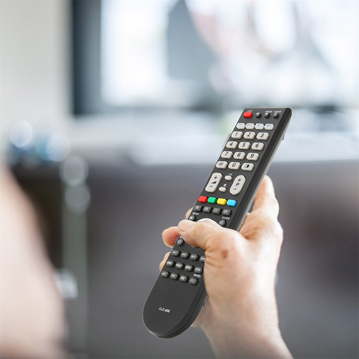 Hand hält eine schwarze Fernbedienung mit mehreren Knöpfen vor einem Fernseher.