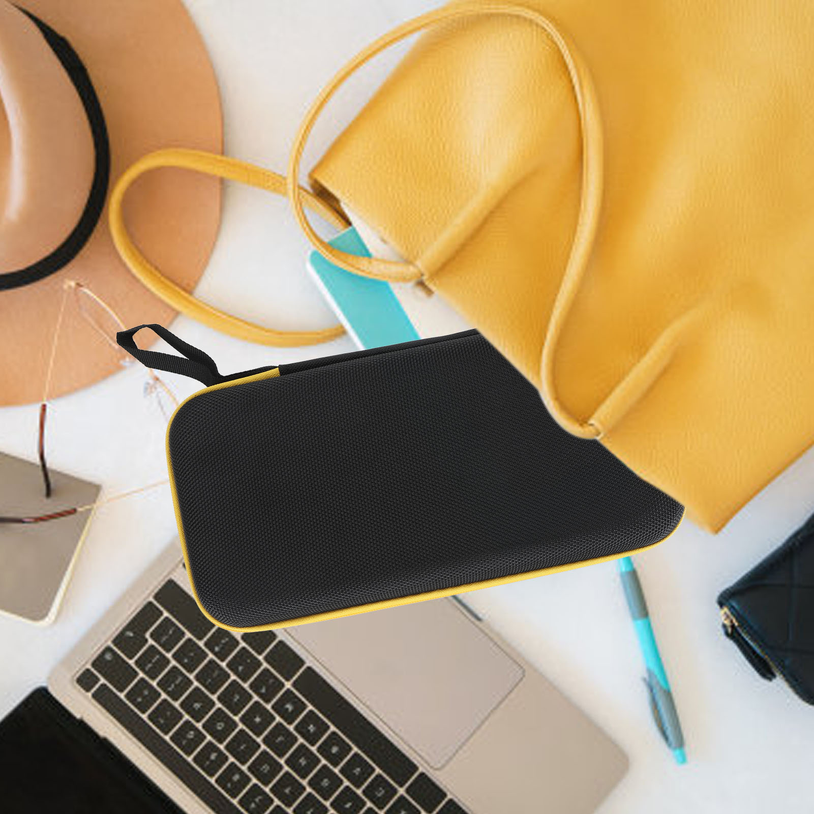 Eine schwarze Hülle neben einer gelben Tasche, einem Hut und einem Laptop auf einer weißen Oberfläche.