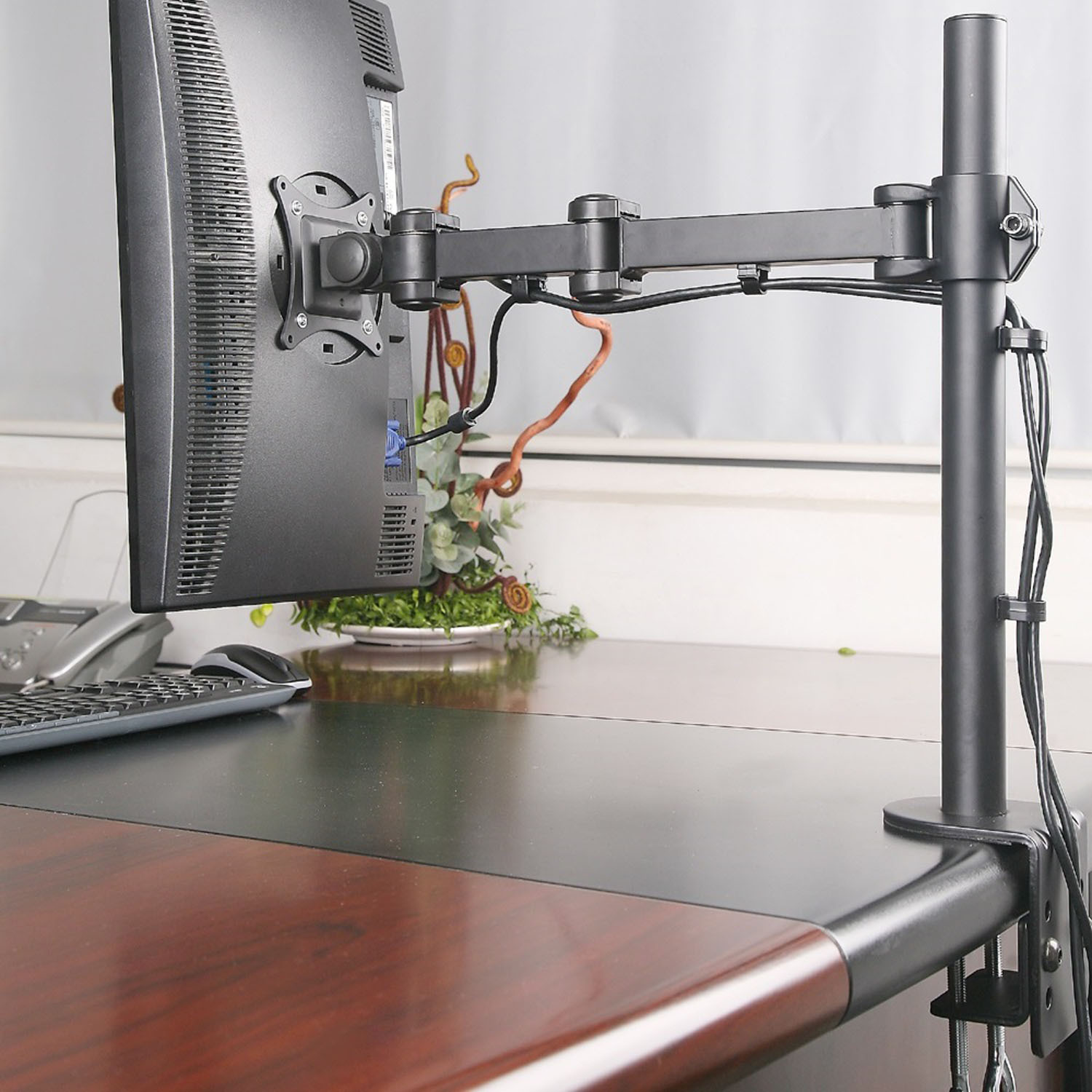 Monitor an einem schwarzen Arm, an einem Holzschreibtisch befestigt. Tastatur und Maus sind sichtbar. Hintergrund ist ein Fenster.