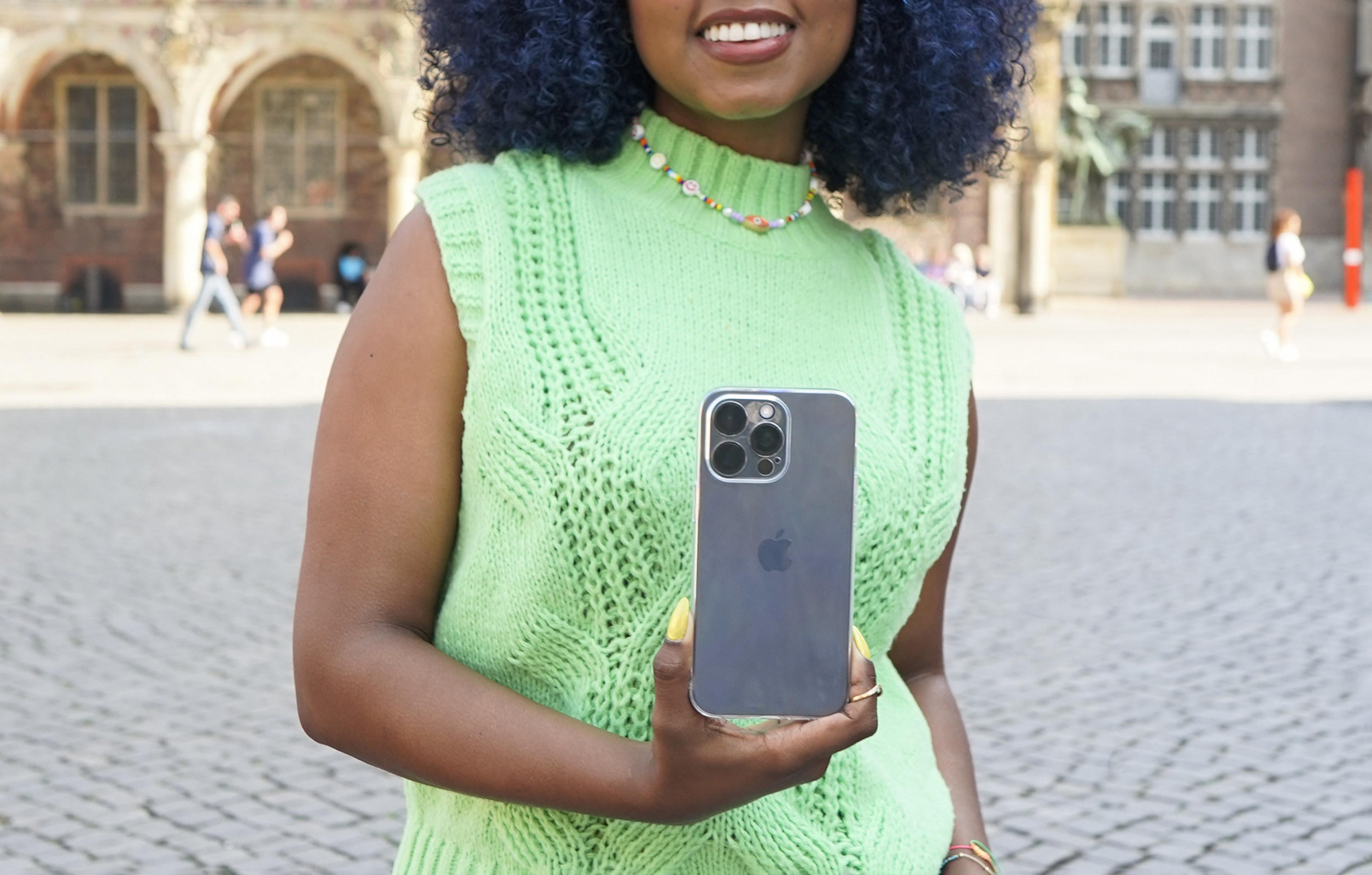 Eine Frau hält ein dunkelgraues Telefon vor einem Backsteingebäude hoch. Sie trägt eine grüne Strickweste.