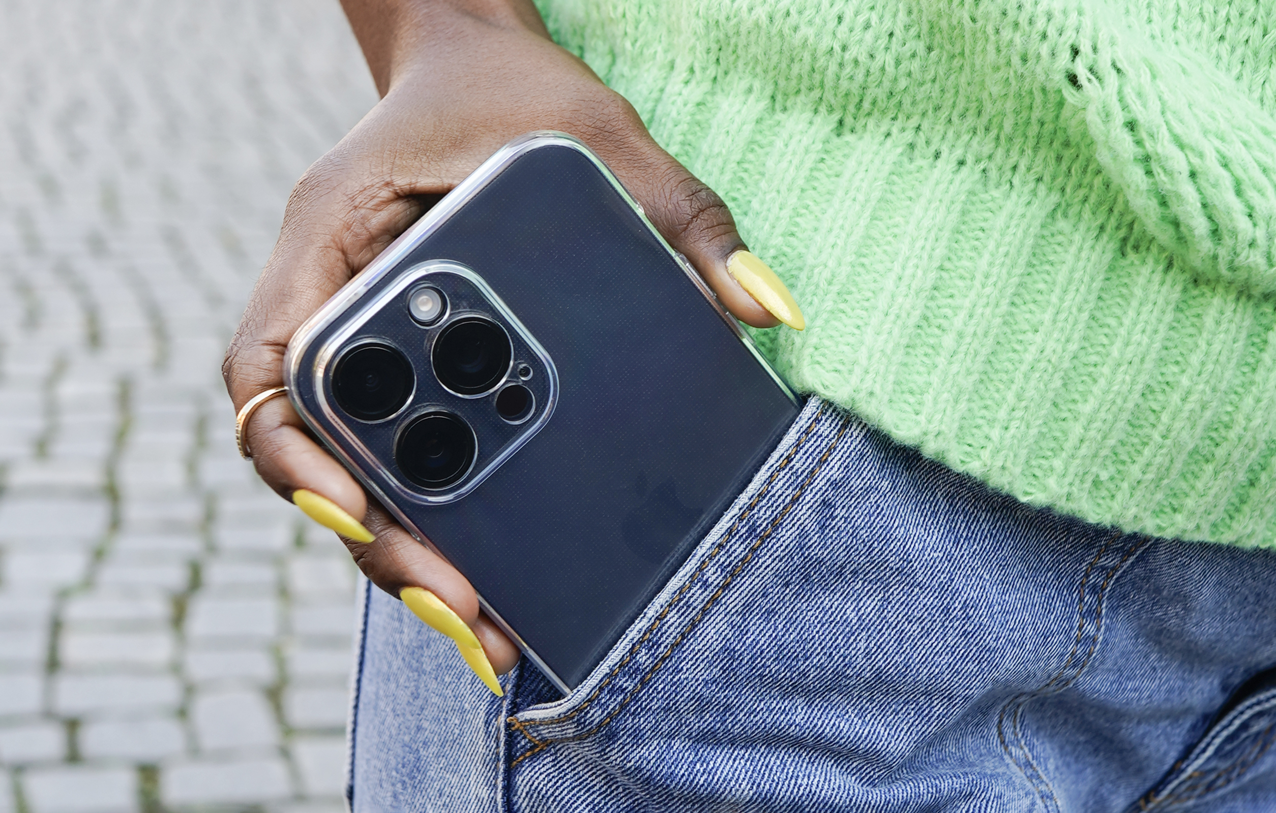 Eine Hand in einem grünen Pullover hält ein Telefon in einer Hülle in einer Jeanshosentasche. Die Nägel sind gelb.