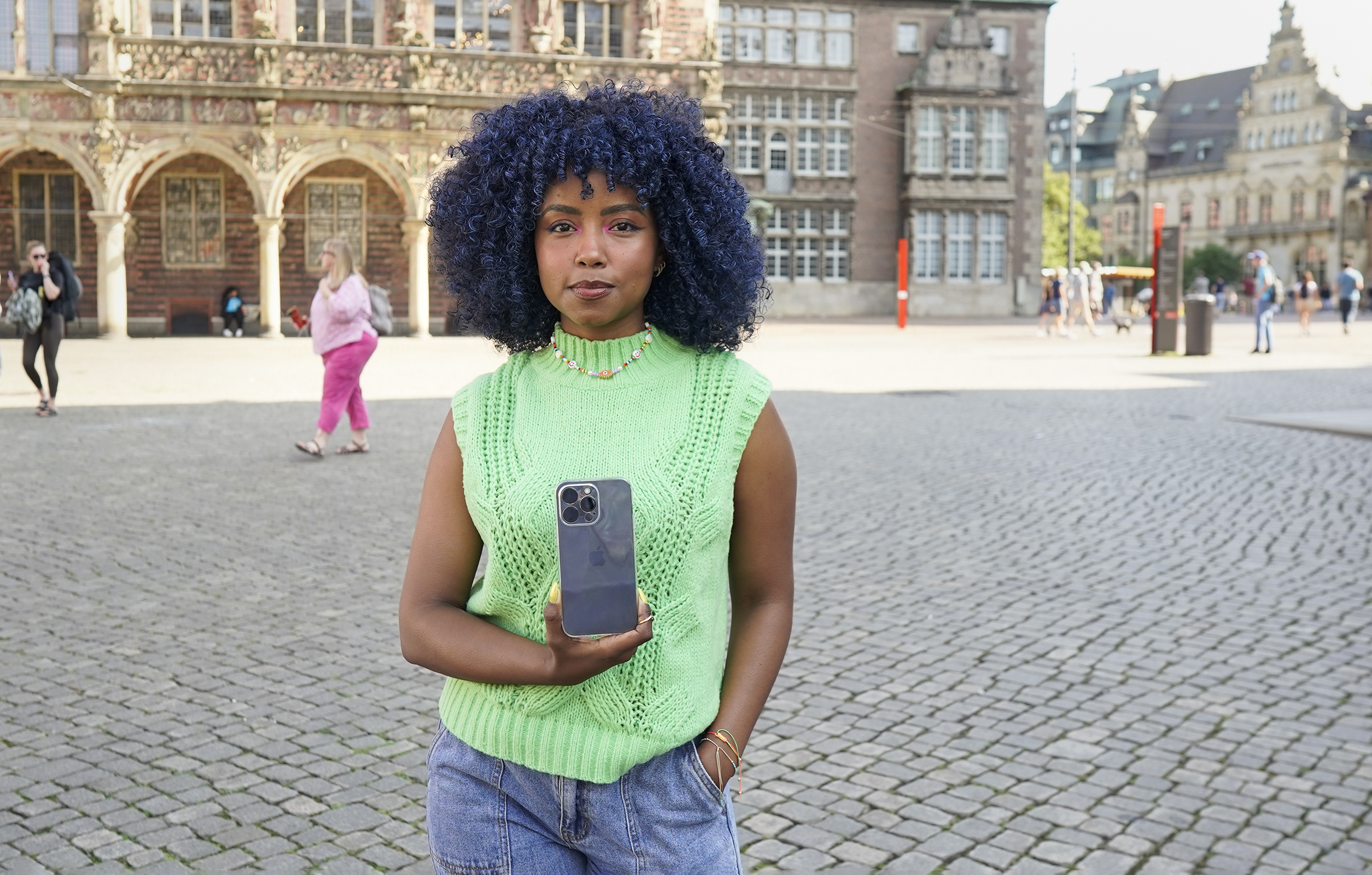 Eine Frau hält ein Telefon vor einem Gebäude. Sie trägt eine grüne Weste und hält das Telefon.