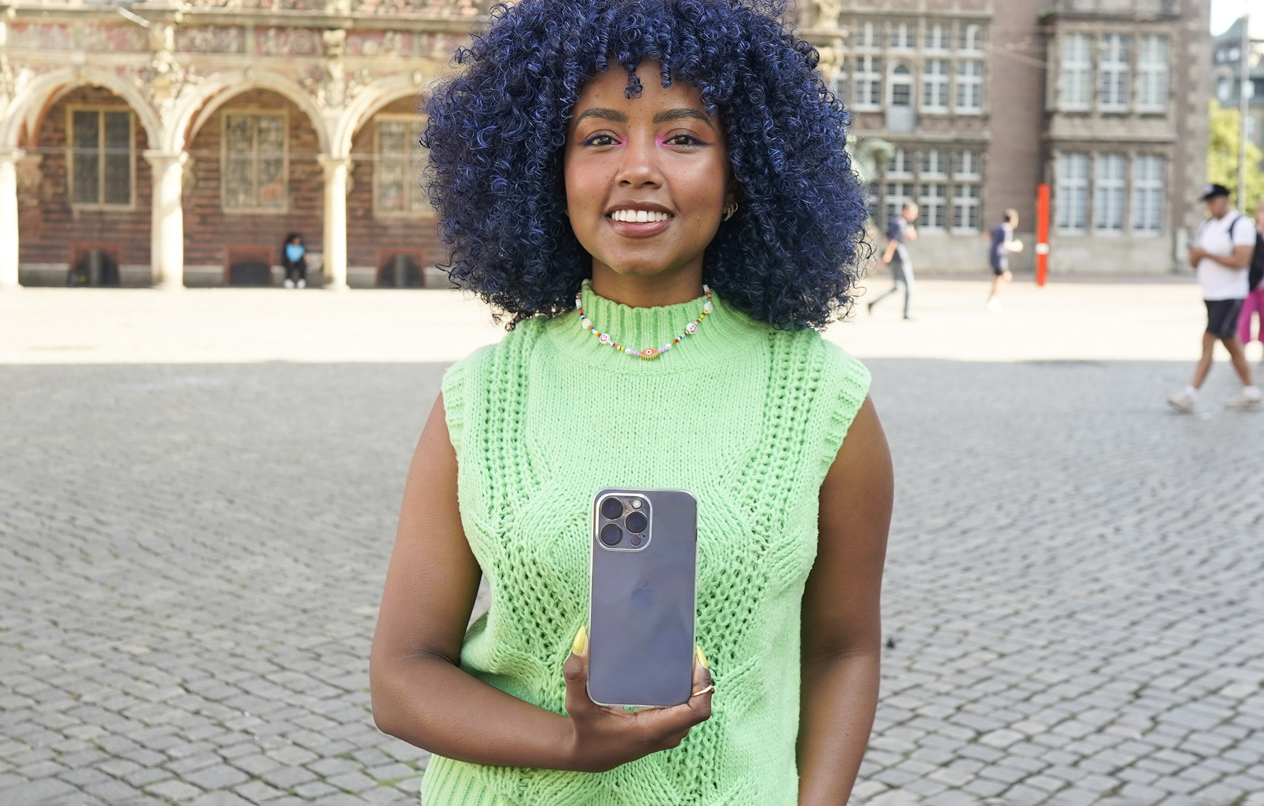Frau mit blauen Haaren, trägt eine grüne Weste, hält ein schwarzes Telefon auf einem Stadtplatz.
