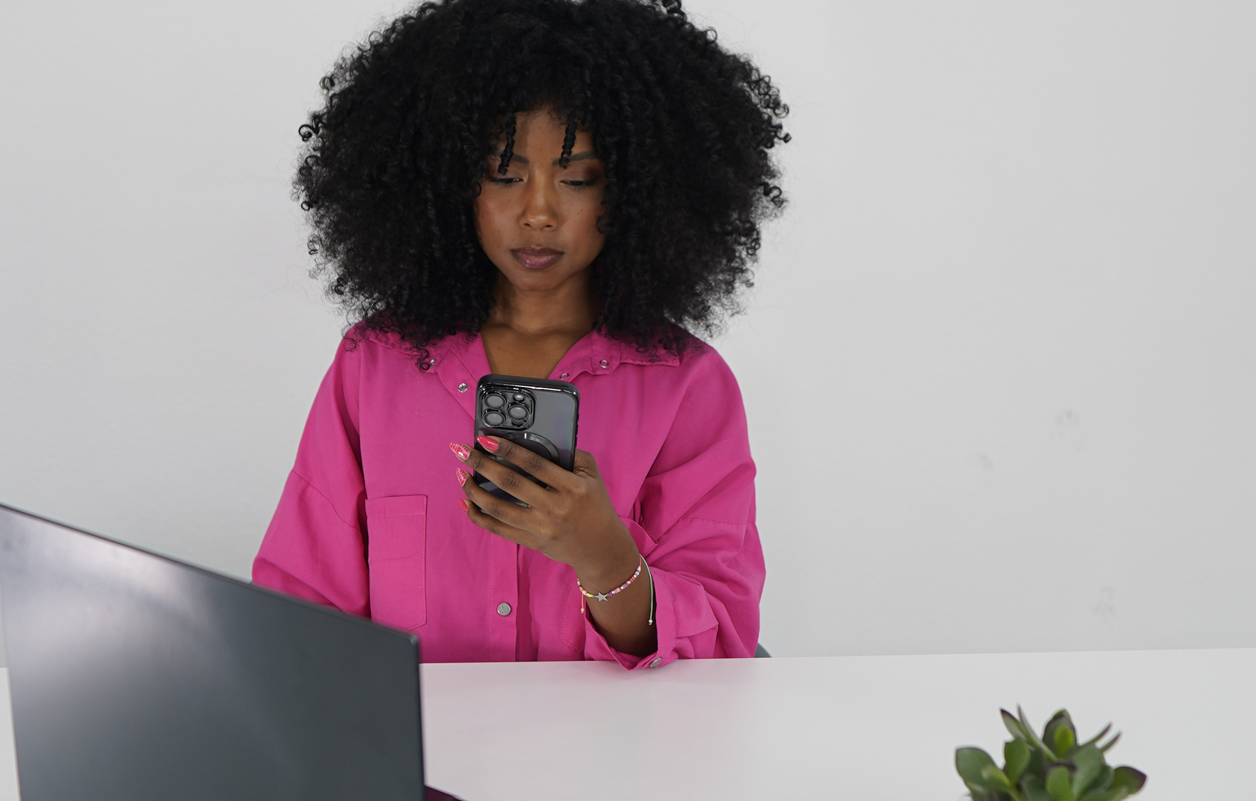 Eine Frau sitzt an einem Schreibtisch und schaut auf ihr Handy. Sie trägt ein rosa Hemd. Ein Laptop und eine Pflanze sind in der Nähe.