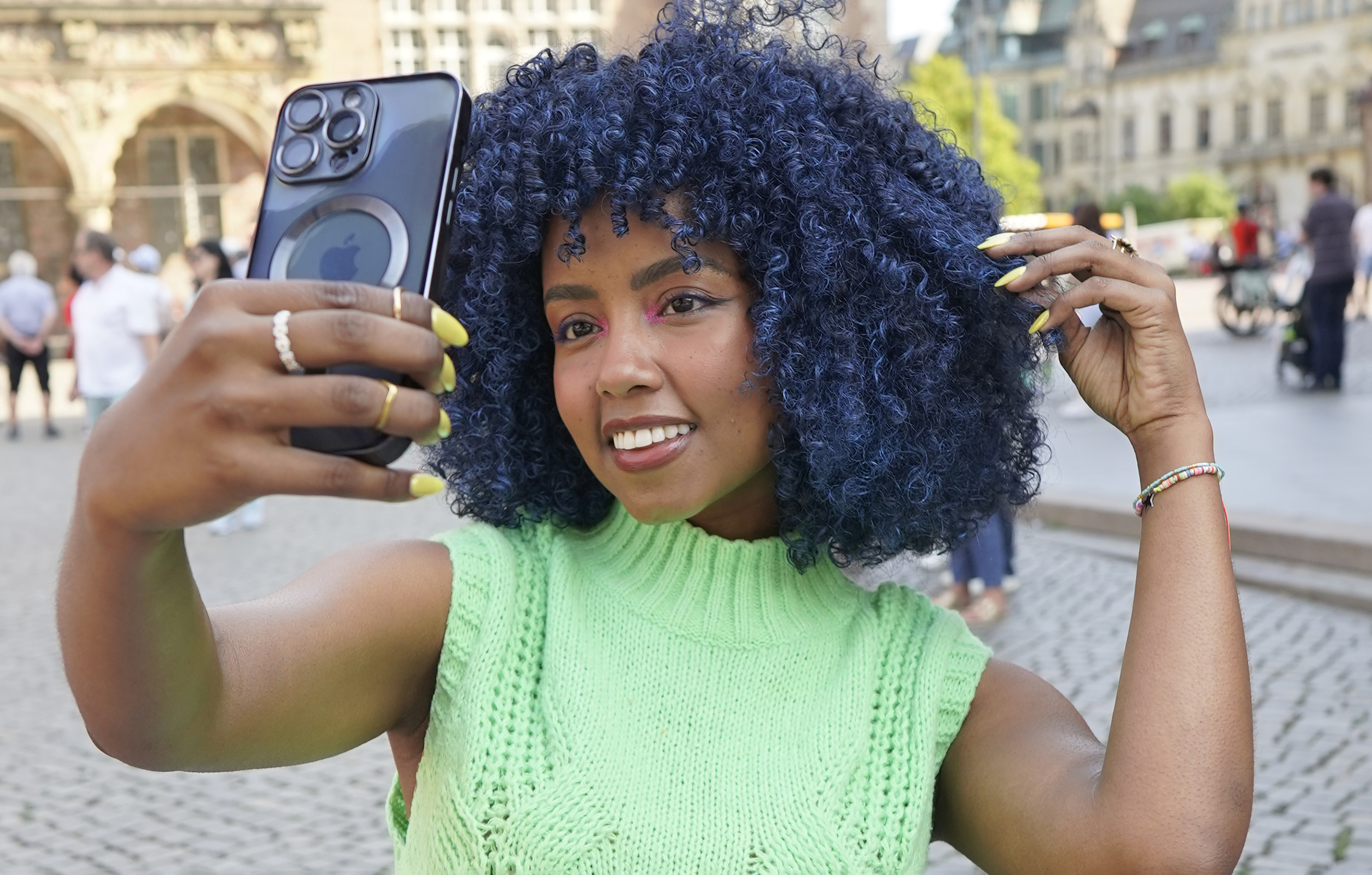Frau mit blauen Locken macht Selfie mit schwarzem Telefon auf Stadtplatz. Grünes Oberteil und rosa Make-up.