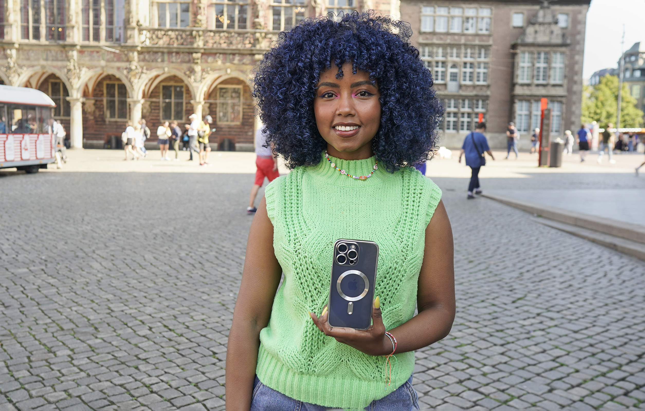 Eine Frau mit blauen Haaren hält ein Telefon vor einem Stadt-Hintergrund. Sie lächelt in die Kamera.