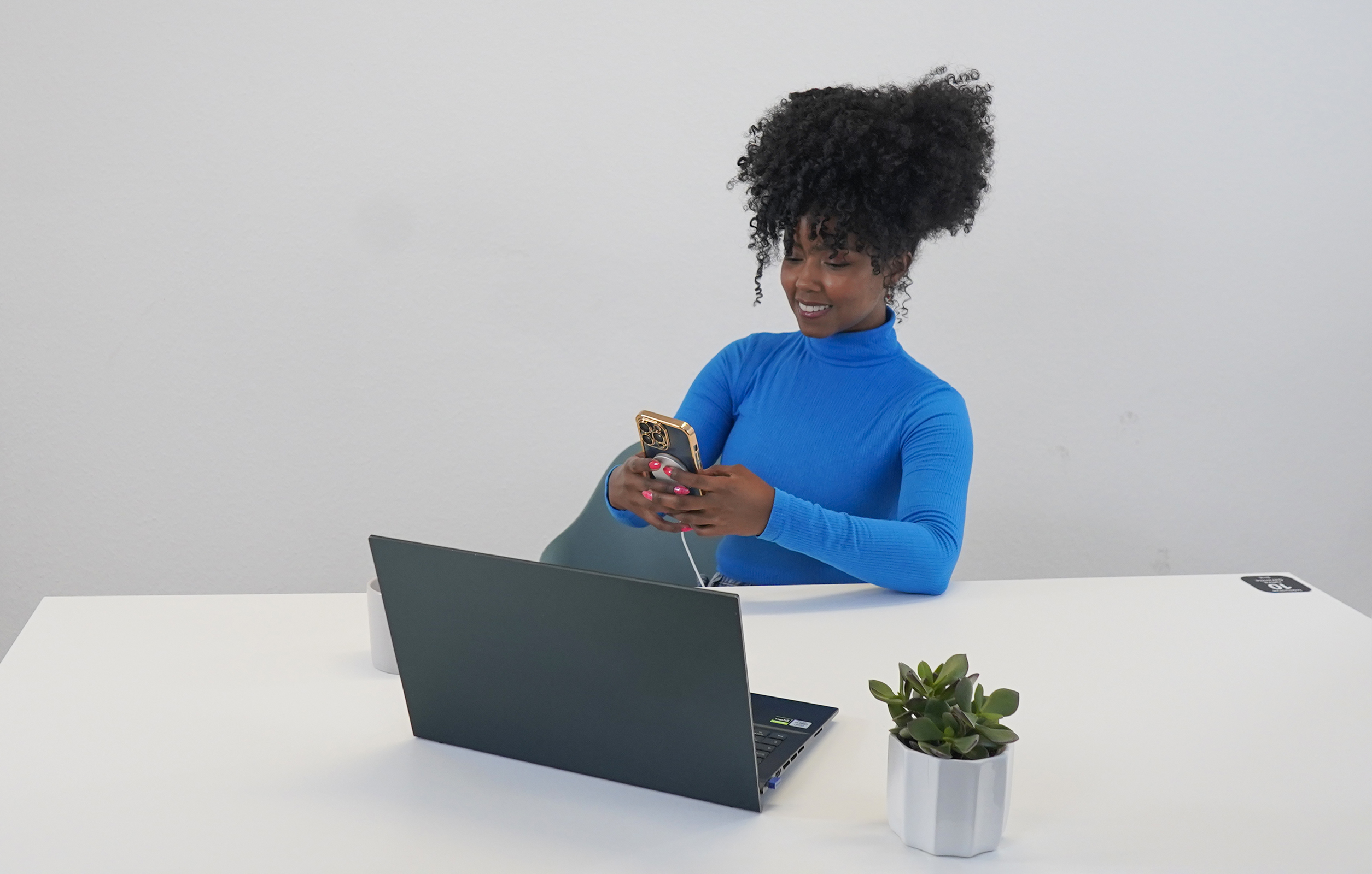 Eine Frau in einem blauen Rollkragen sitzt an einem Schreibtisch und benutzt ein Telefon und einen Laptop, daneben eine Pflanze.