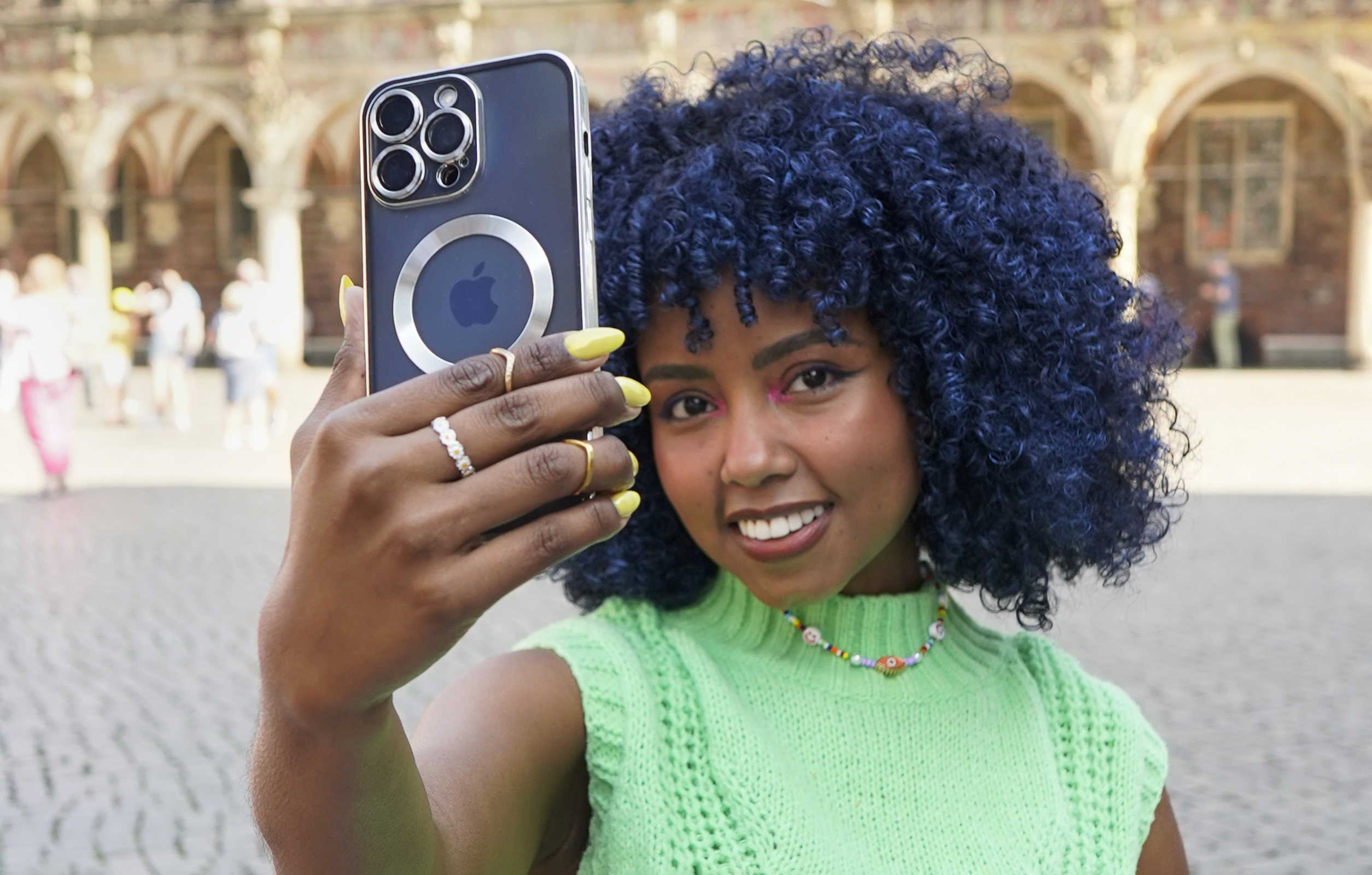Frau mit blauen Locken macht ein Selfie mit einem Telefon. Sie trägt ein grünes Oberteil und eine Halskette.