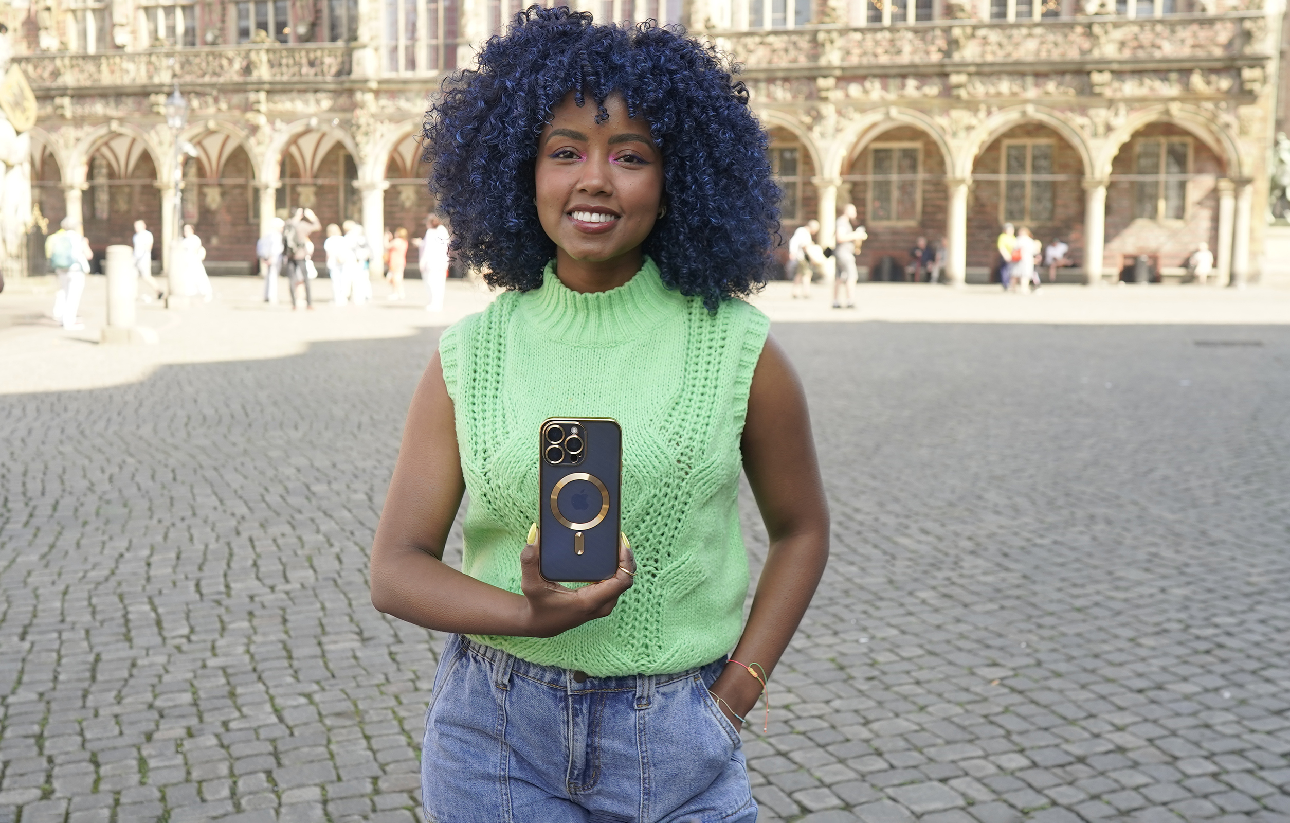 Frau mit blauen Locken hält eine Handyhülle auf einem Stadtplatz.