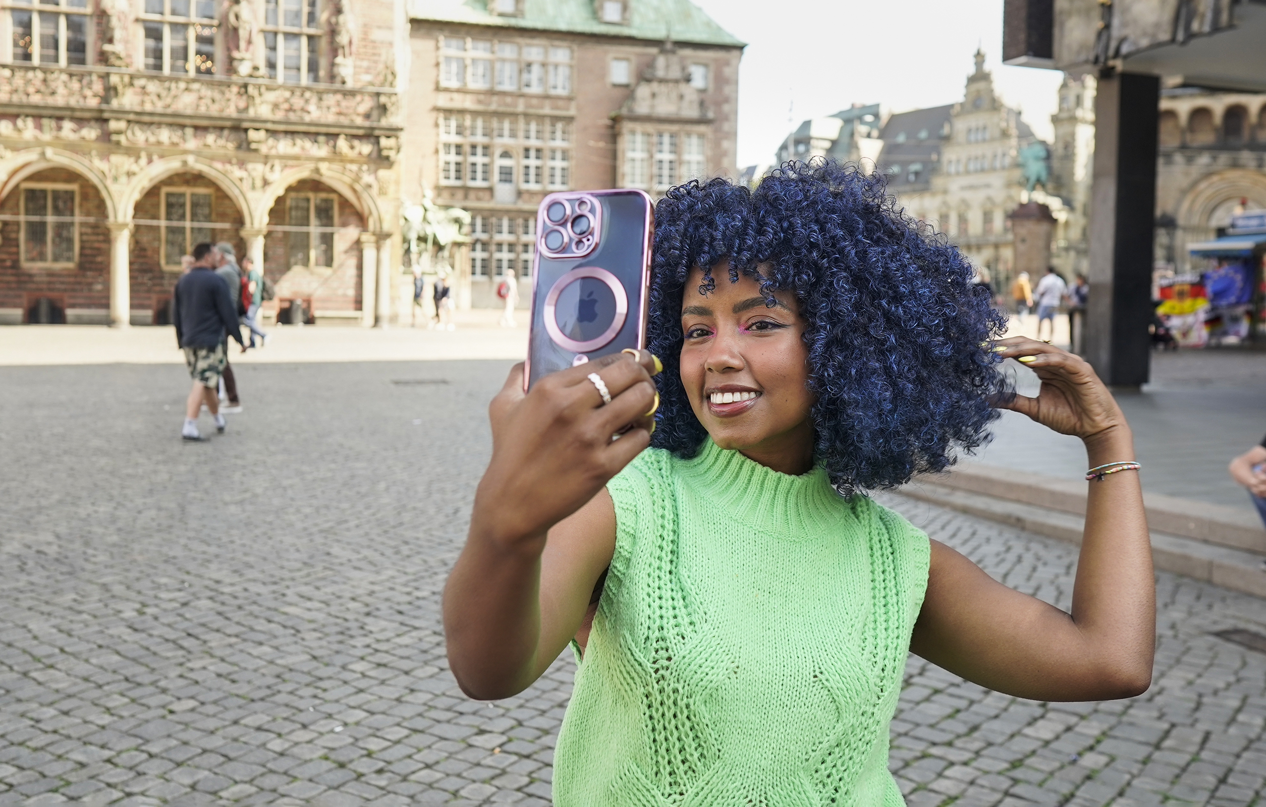 Eine Frau macht ein Selfie auf einem Stadtplatz, hält ein Telefon mit rosa Hülle.
