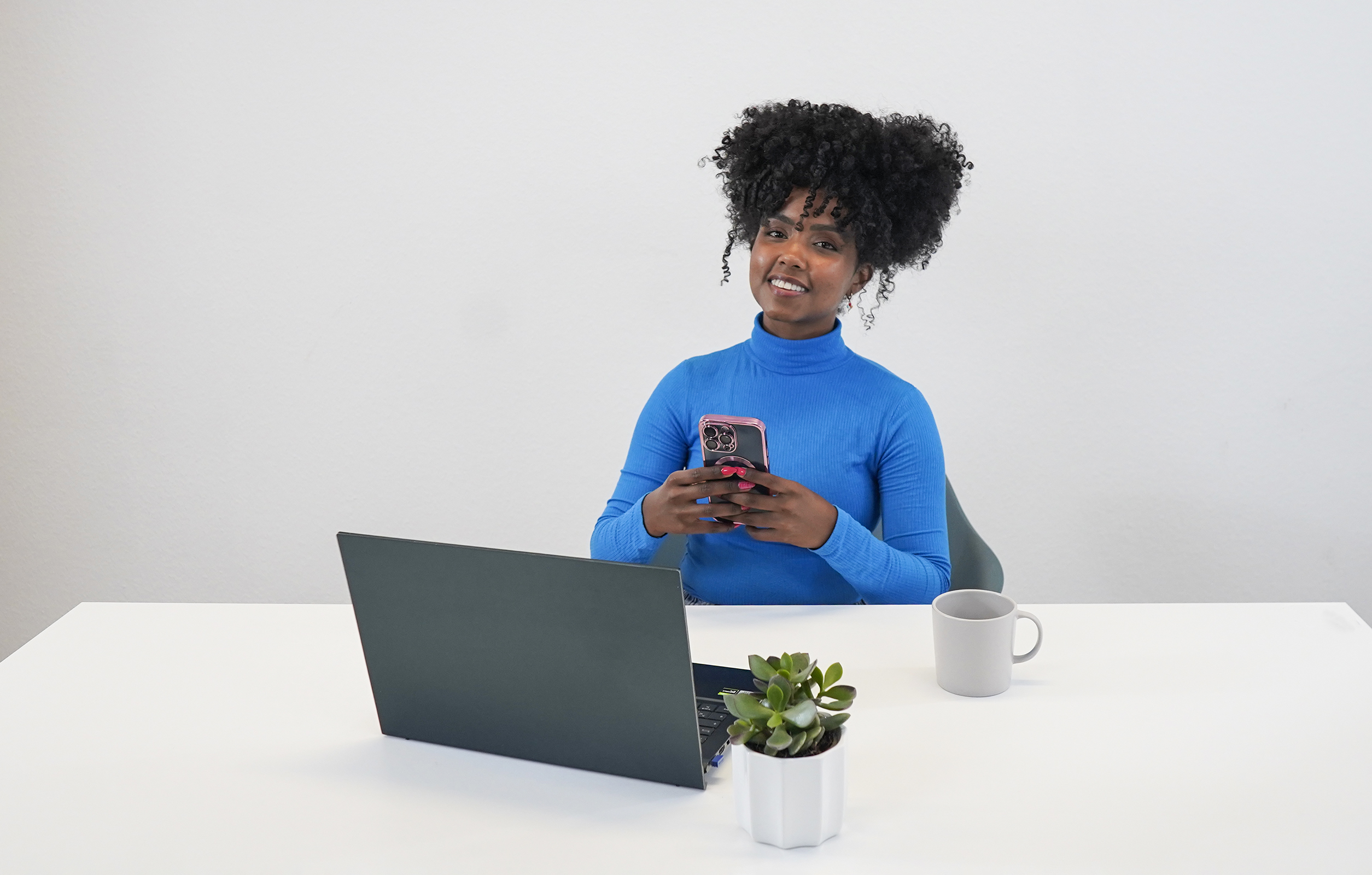 Eine Frau mit lockigem Haar benutzt ein Telefon an einem Schreibtisch mit Laptop und Pflanze.