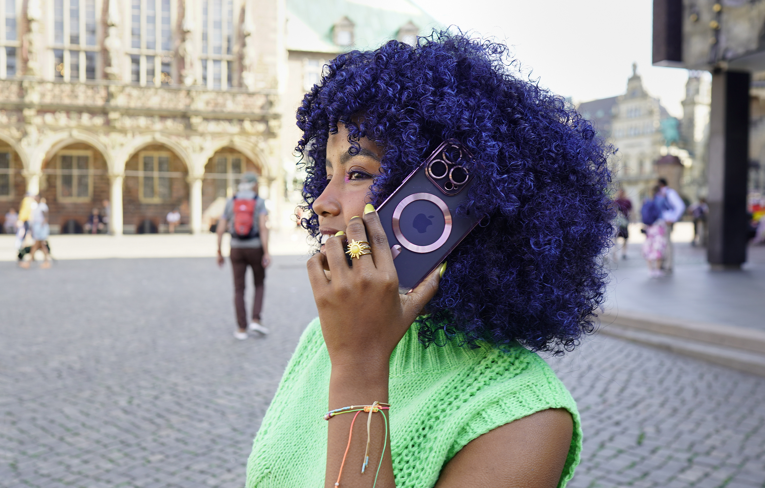 Eine Frau hält ein Telefon an ihr Ohr. Sie hat blaue Locken. Ein Backsteingebäude ist im Hintergrund.