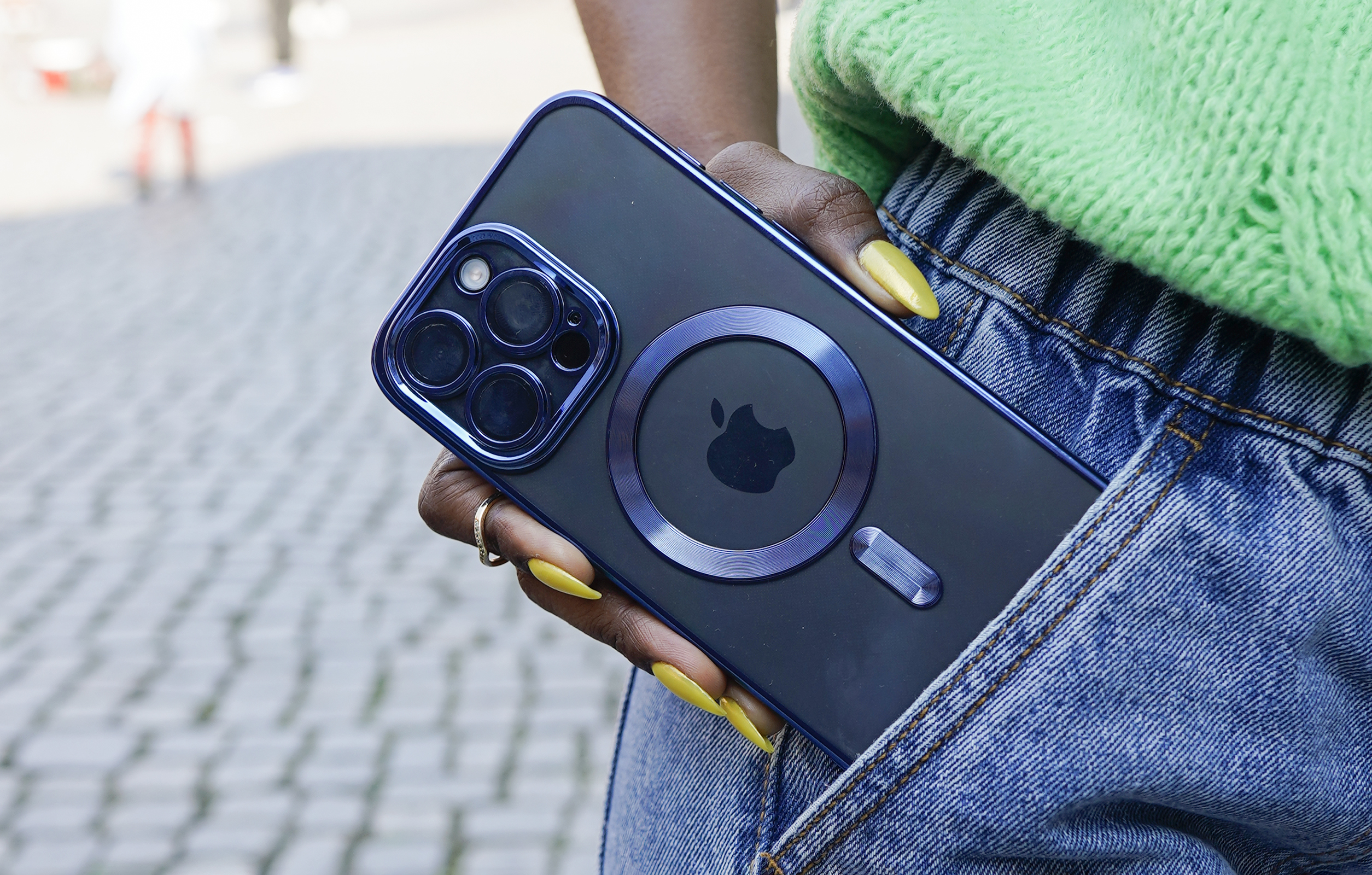 Eine Person hält ein Telefon mit Hülle, trägt eine Jeansjacke und einen grünen Pullover.