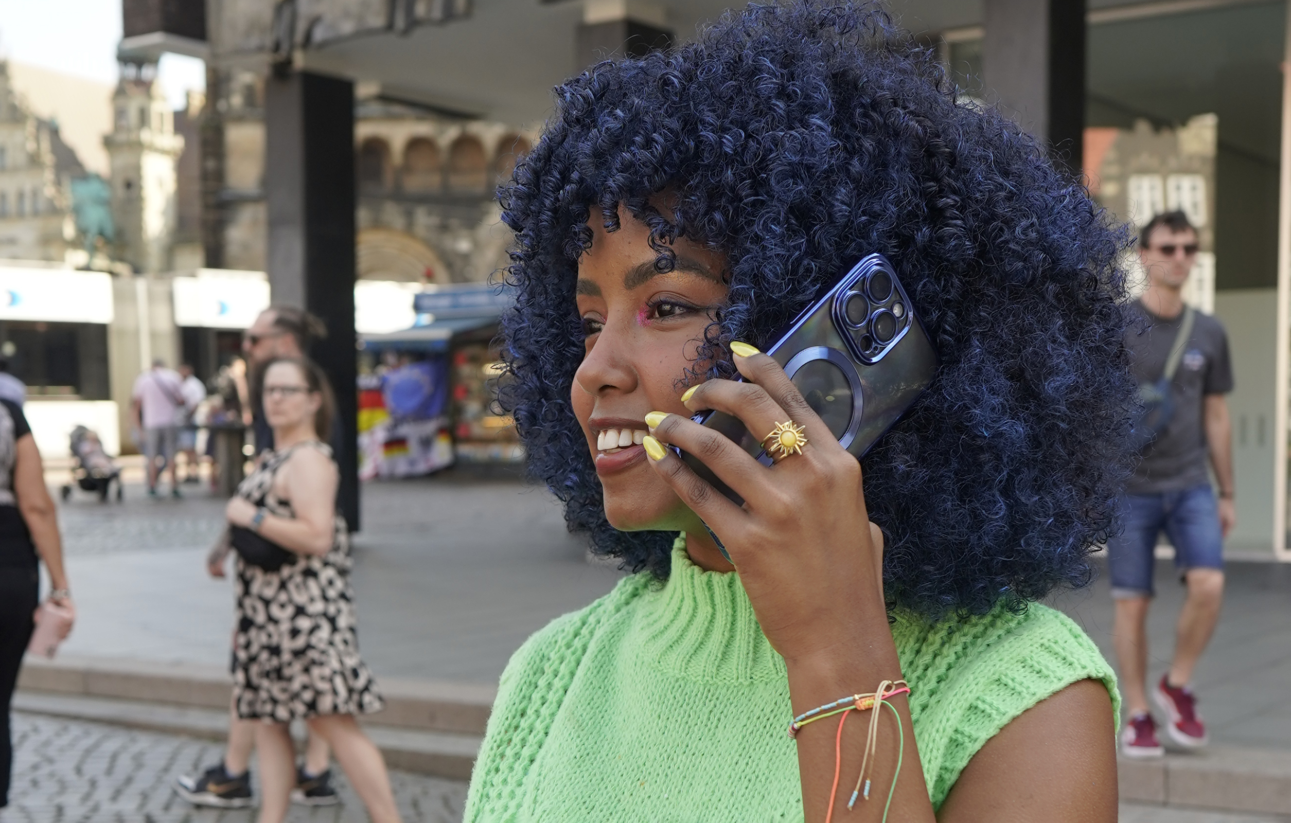 Eine Frau mit blauen Haaren telefoniert und lächelt im Freien.