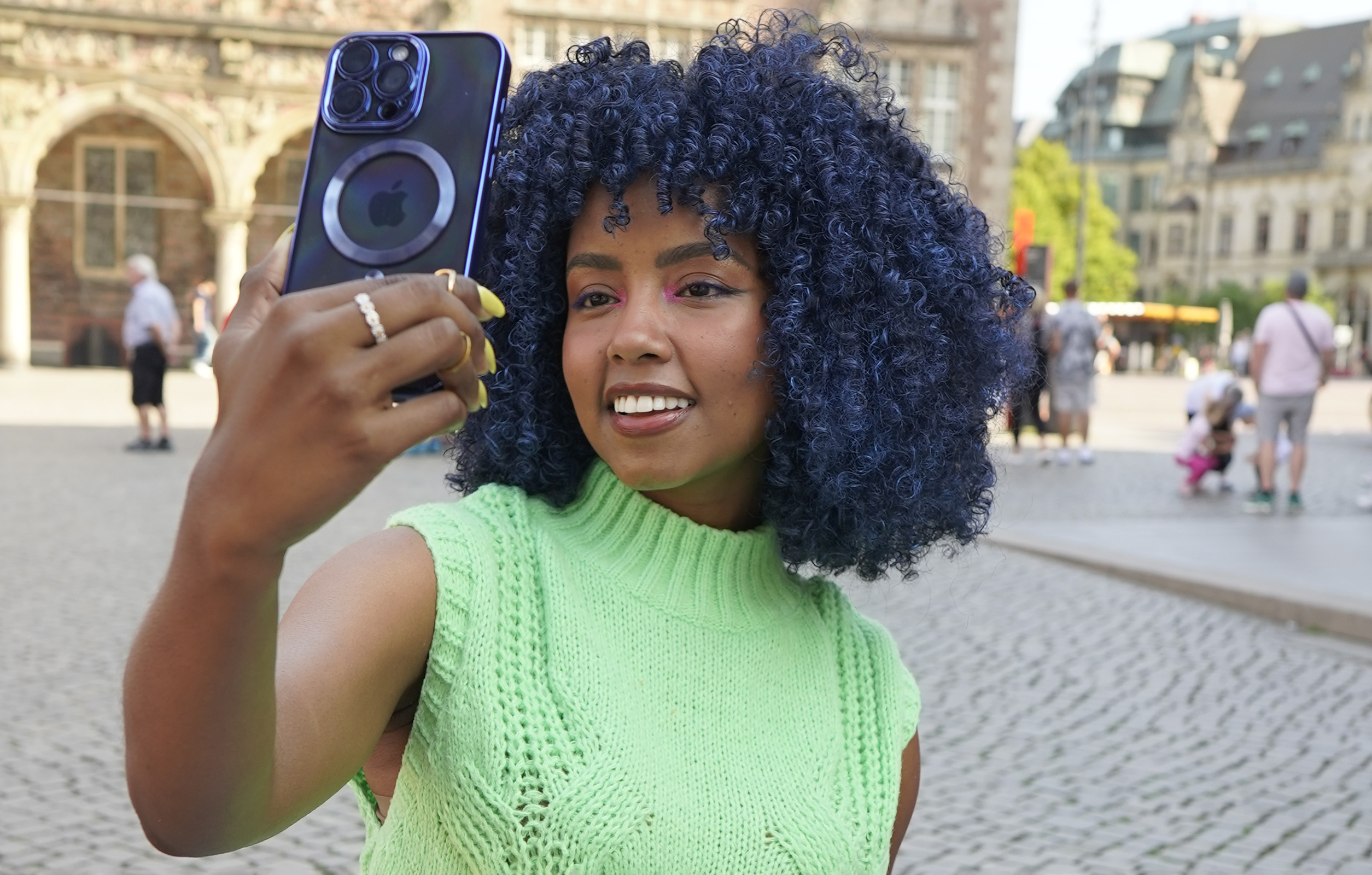 Frau mit blauen Locken macht Selfie mit Handy. Sie trägt grünes Oberteil und steht draußen.
