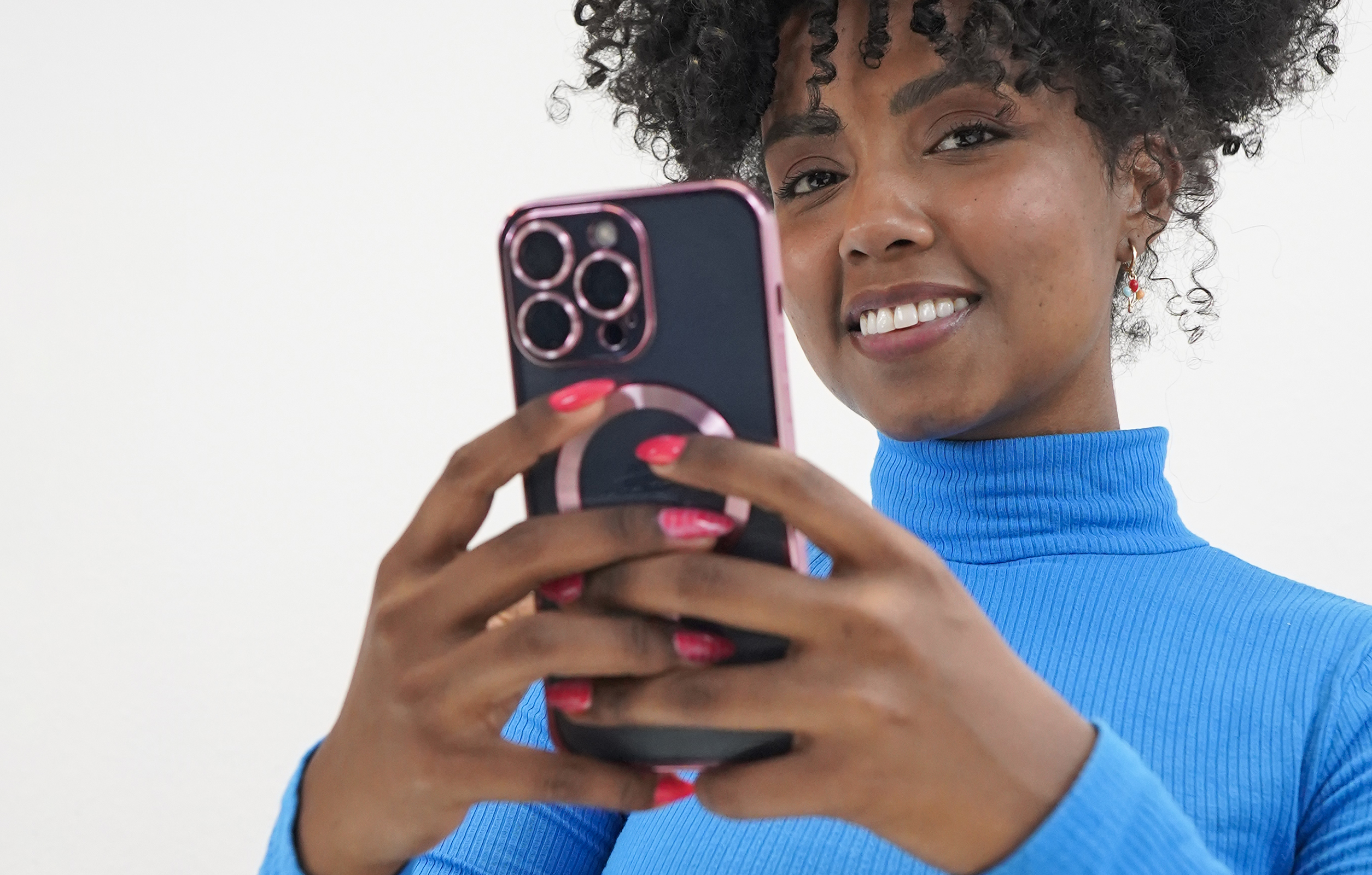 Eine lächelnde Frau in einem blauen Rollkragenpullover, die ein Telefon mit schwarzem Case und pinkem Popsocket hält.