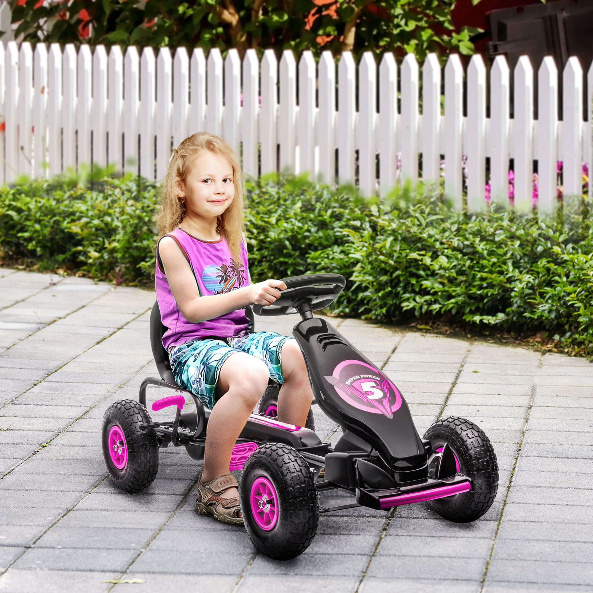 Una bambina guida un kart a pedali rosa e nero. Sorride, indossa una canotta e pantaloncini.