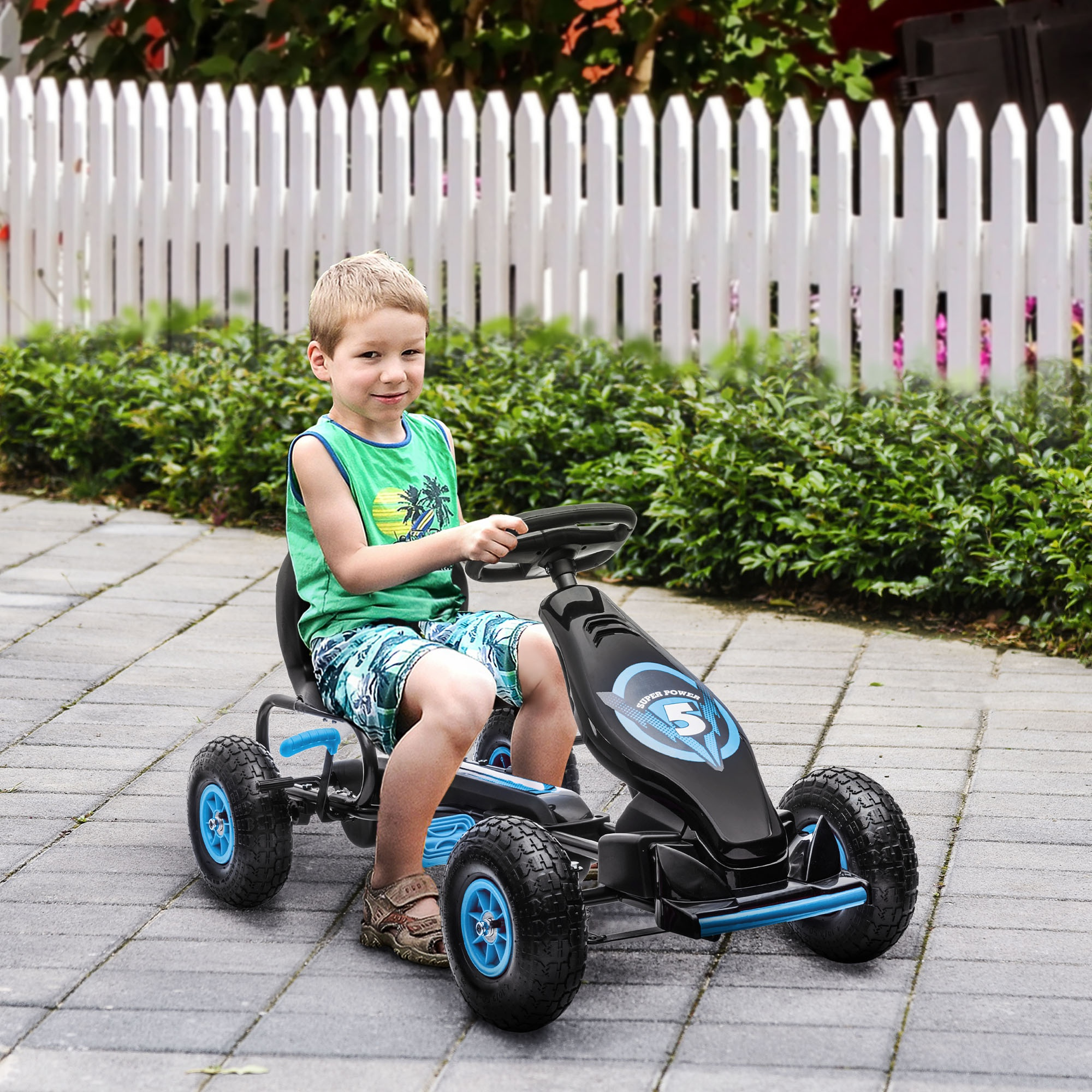 Niño en un kart de pedales, sonriendo. Valla blanca y arbustos verdes al fondo.