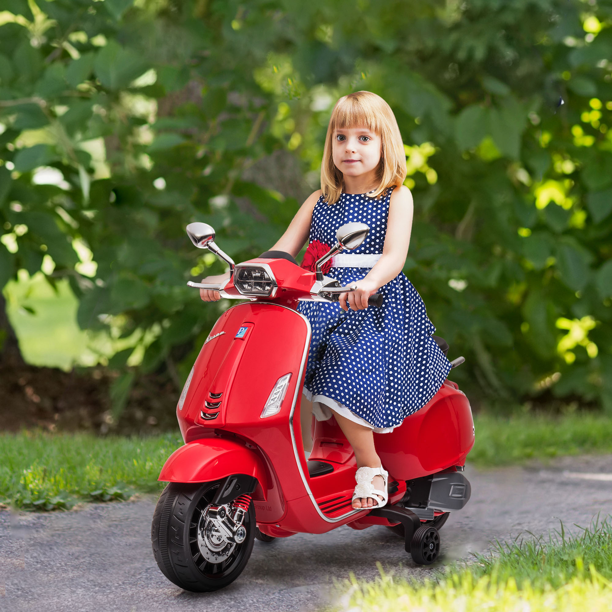 Una niña pequeña monta un scooter de juguete rojo estilo Vespa en un camino pavimentado, rodeada de árboles.