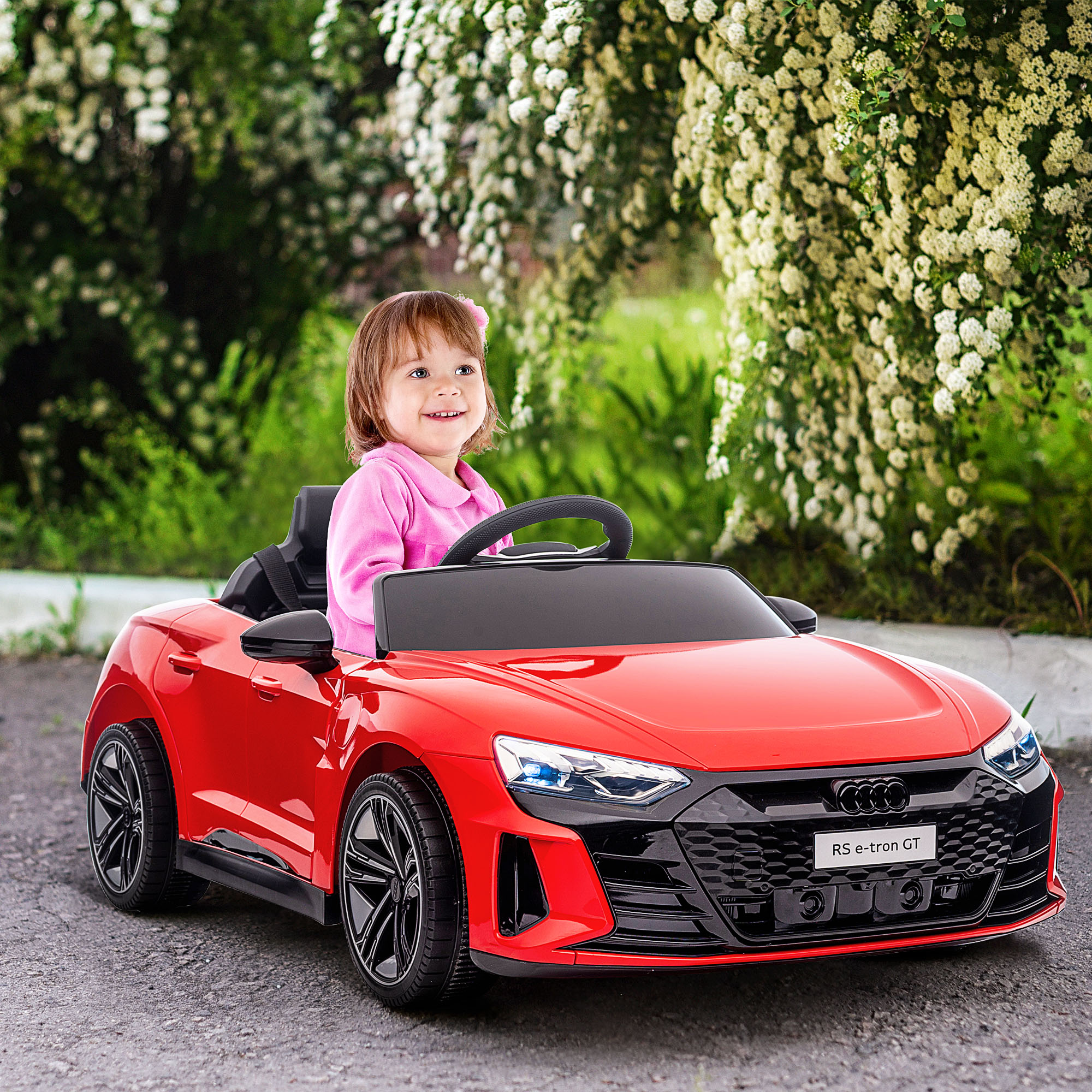 Una bambina sorridente in un'auto giocattolo rossa, che guida su una strada asfaltata con sfondo verde.
