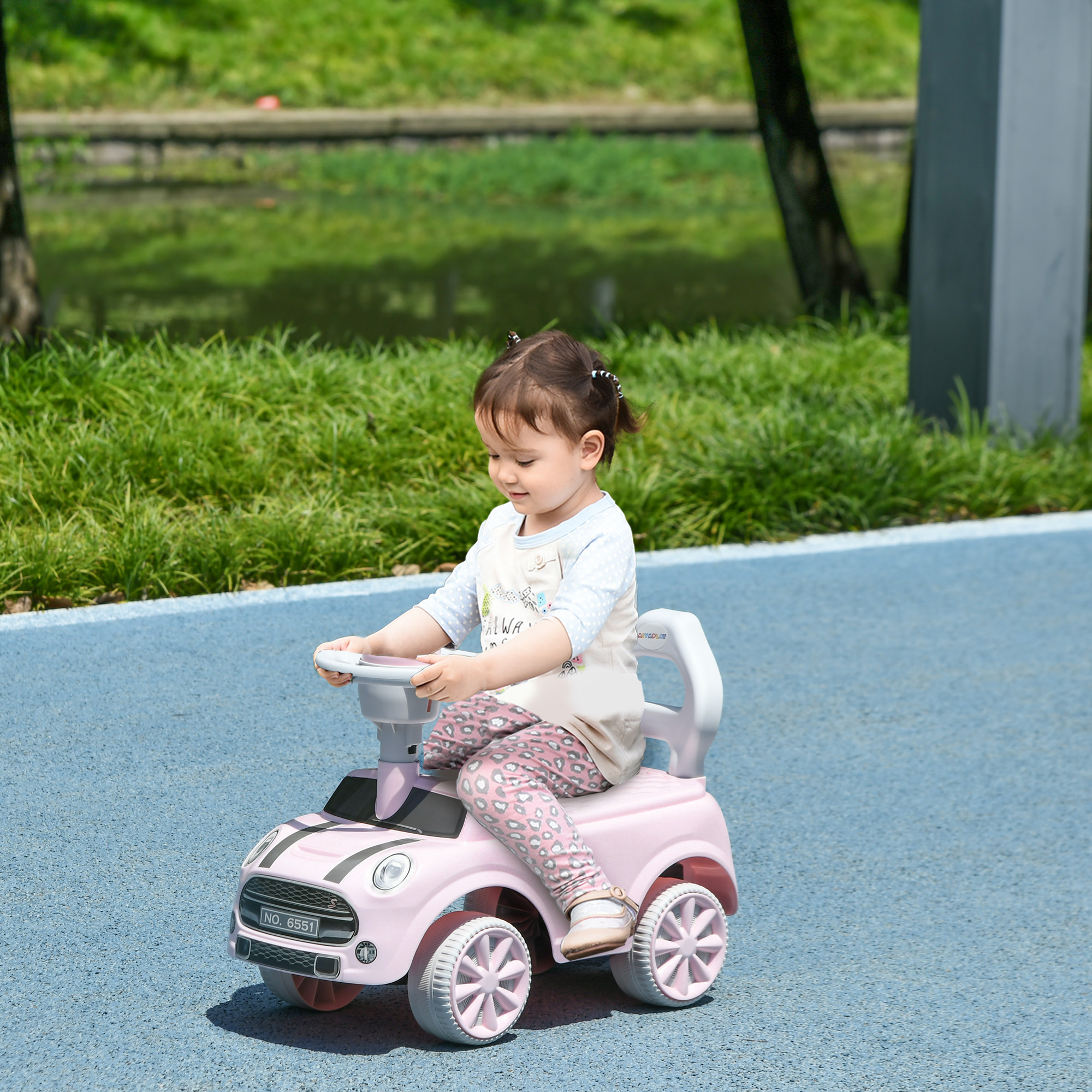 Una niña pequeña está sentada en un coche de juguete rosa. Sonríe. El fondo muestra césped, árboles y un camino azul.