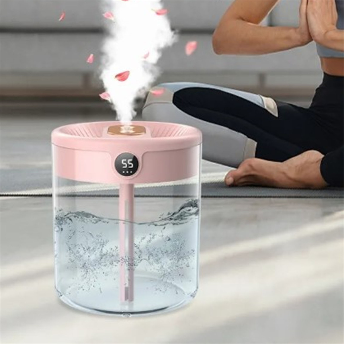 Ein rosa Luftbefeuchter mit Digitalanzeige, der Dampf abgibt. Im Hintergrund eine Person beim Yoga.