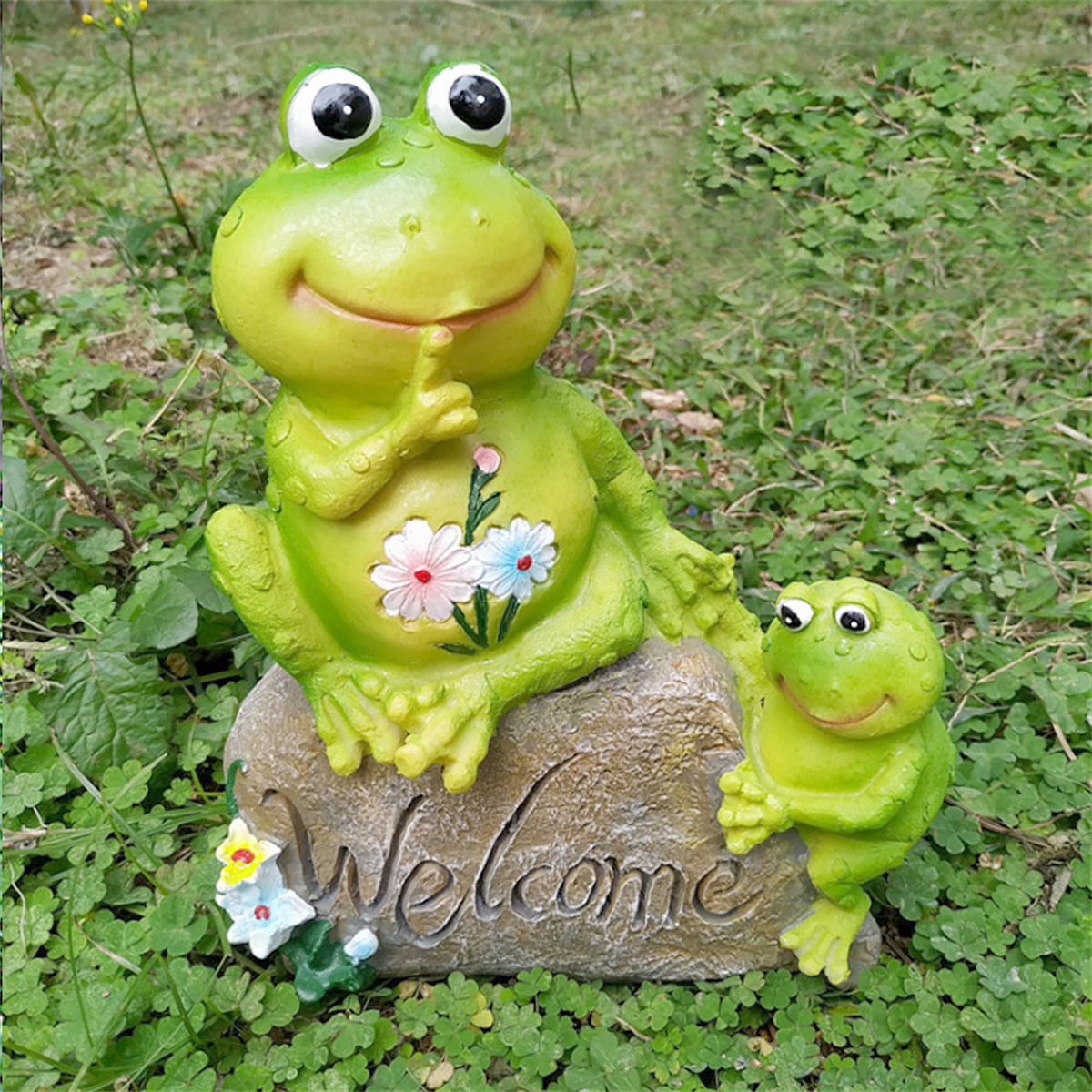 Zwei grüne Frosch-Statuen, eine große, eine kleine, sitzen auf einem Stein mit einem Schild "Willkommen".