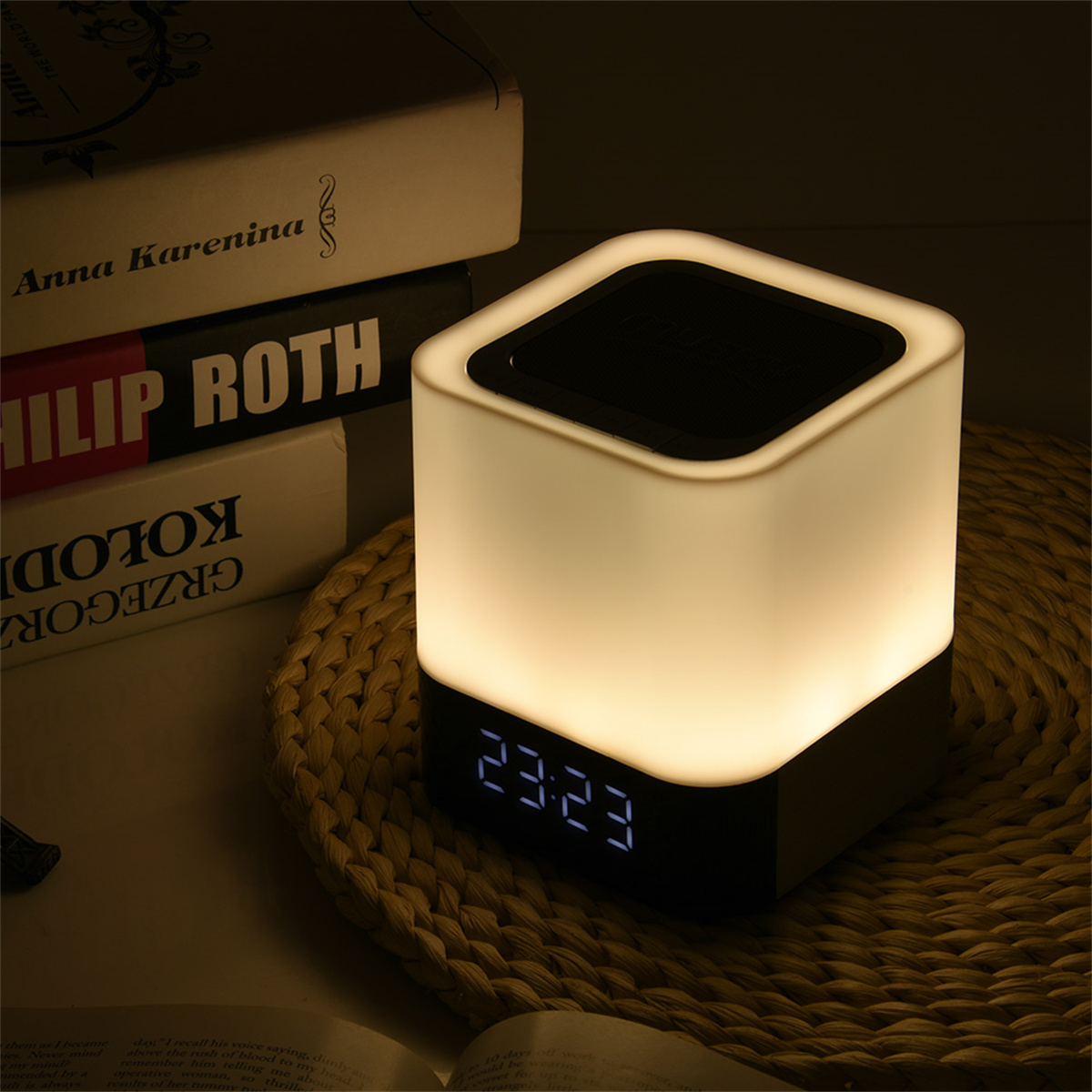 Eine würfelförmige Lampe mit Digitaluhr auf einer gewebten Matte. Bücher sind im Hintergrund.