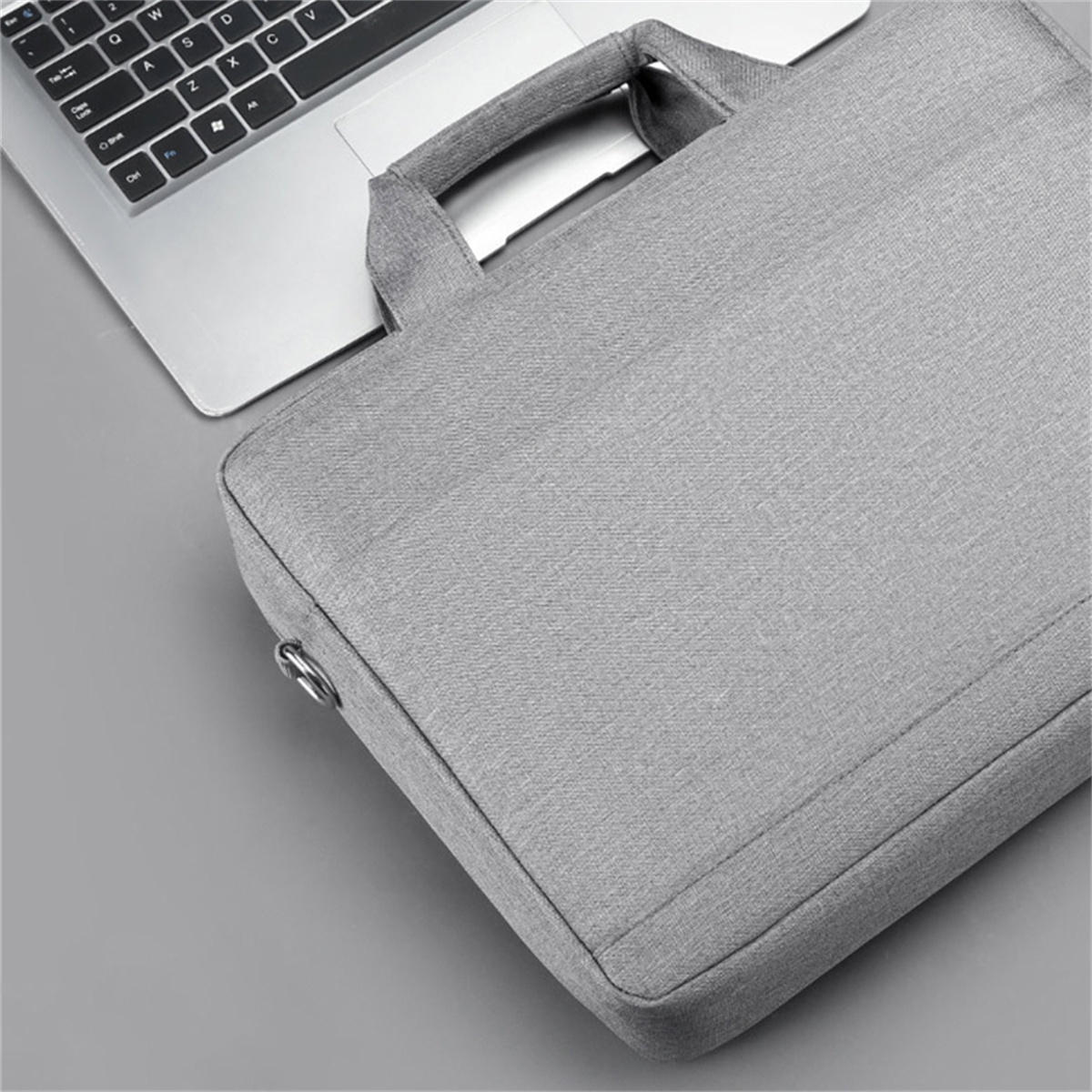 Eine graue Laptoptasche neben einem Laptop auf einer grauen Oberfläche. Die Tasche hat einen Griff.
