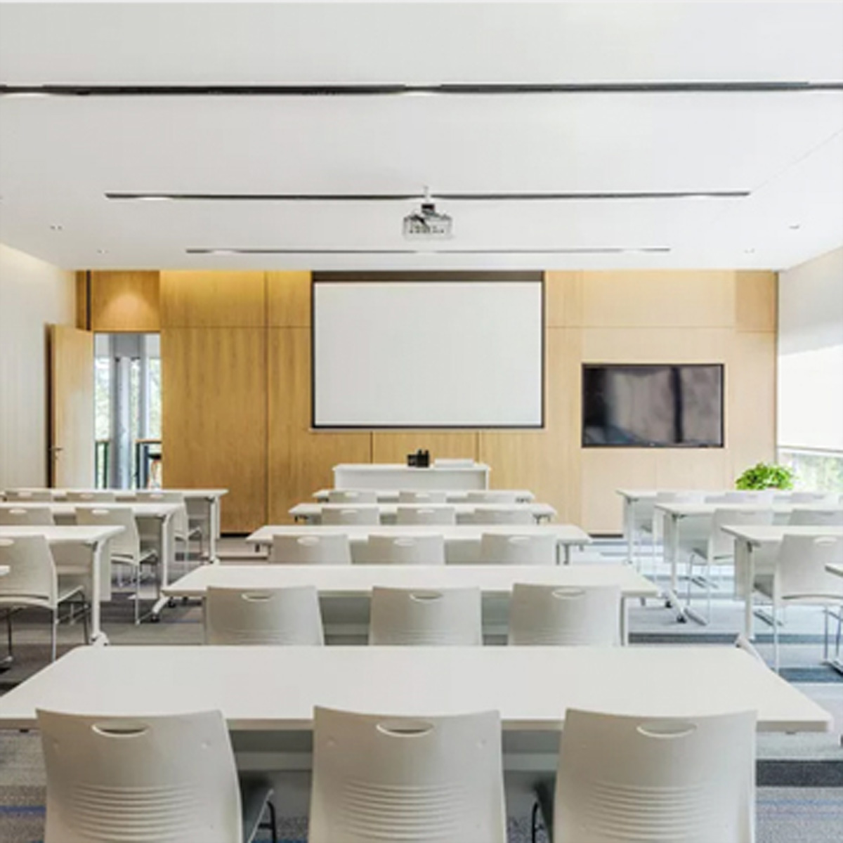 Sala konferencyjna z rzędami białych stołów i krzeseł. Projektor i ekran z przodu. Przestrzeń jest dobrze oświetlona.