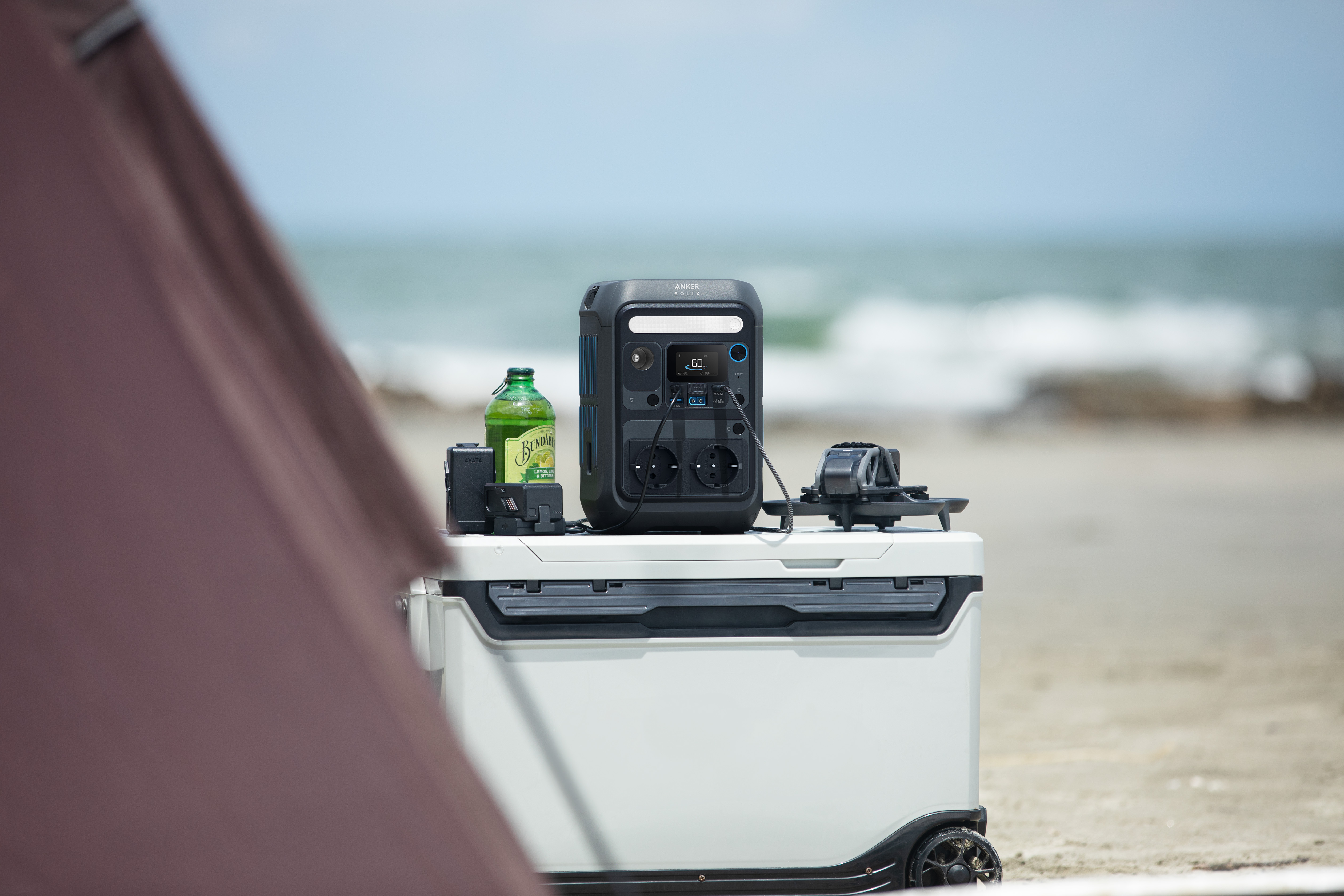 Przenośna stacja zasilania, butelka piwa i inne przedmioty na lodówce na plaży.