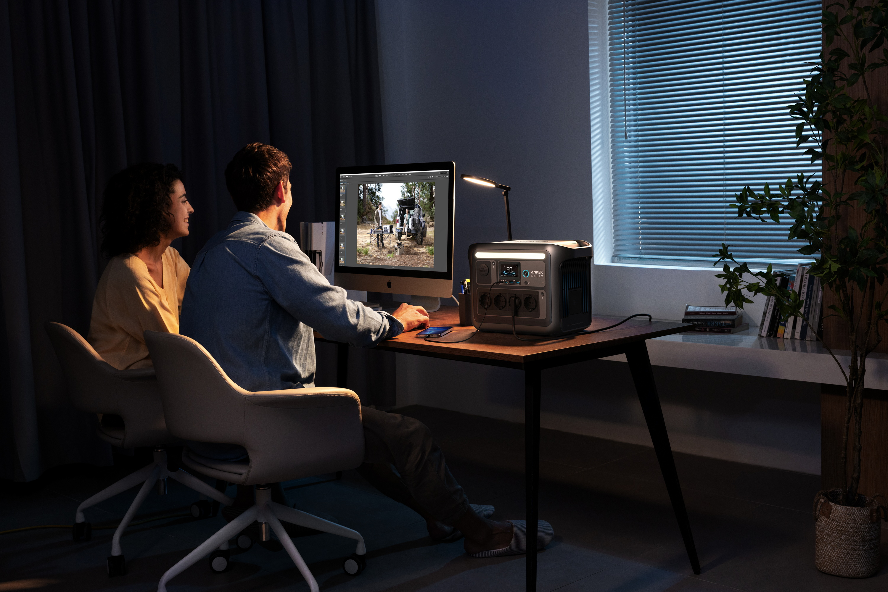 Para w ciemnym pokoju pracuje przy biurku z komputerem. Przenośna stacja zasilania na biurku.