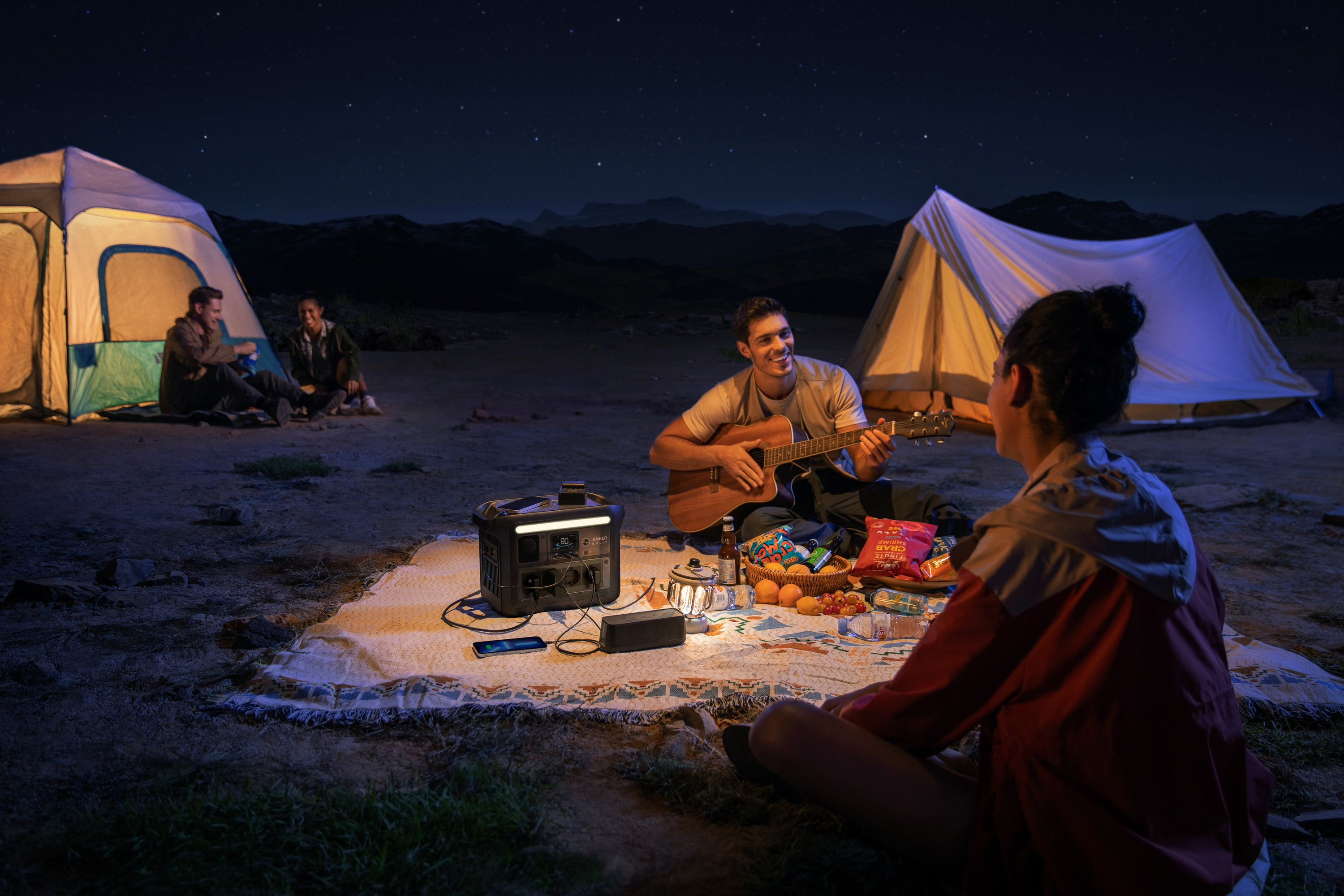 Grupa osób biwakujących w nocy z gitarą i przenośną stacją zasilania.