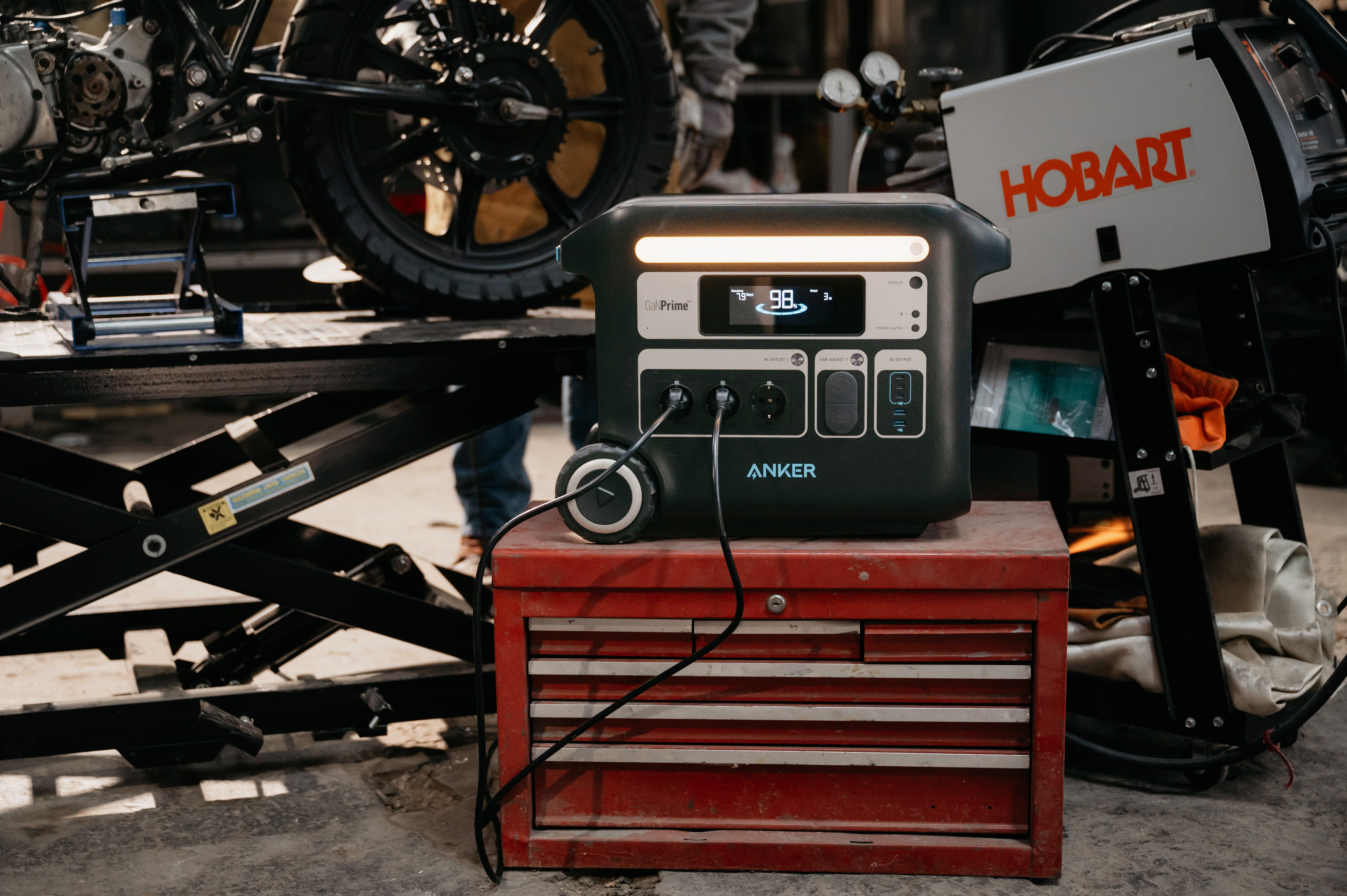 Czarna stacja zasilania Anker na czerwonej skrzynce narzędziowej. Motocykl i spawarka w tle.