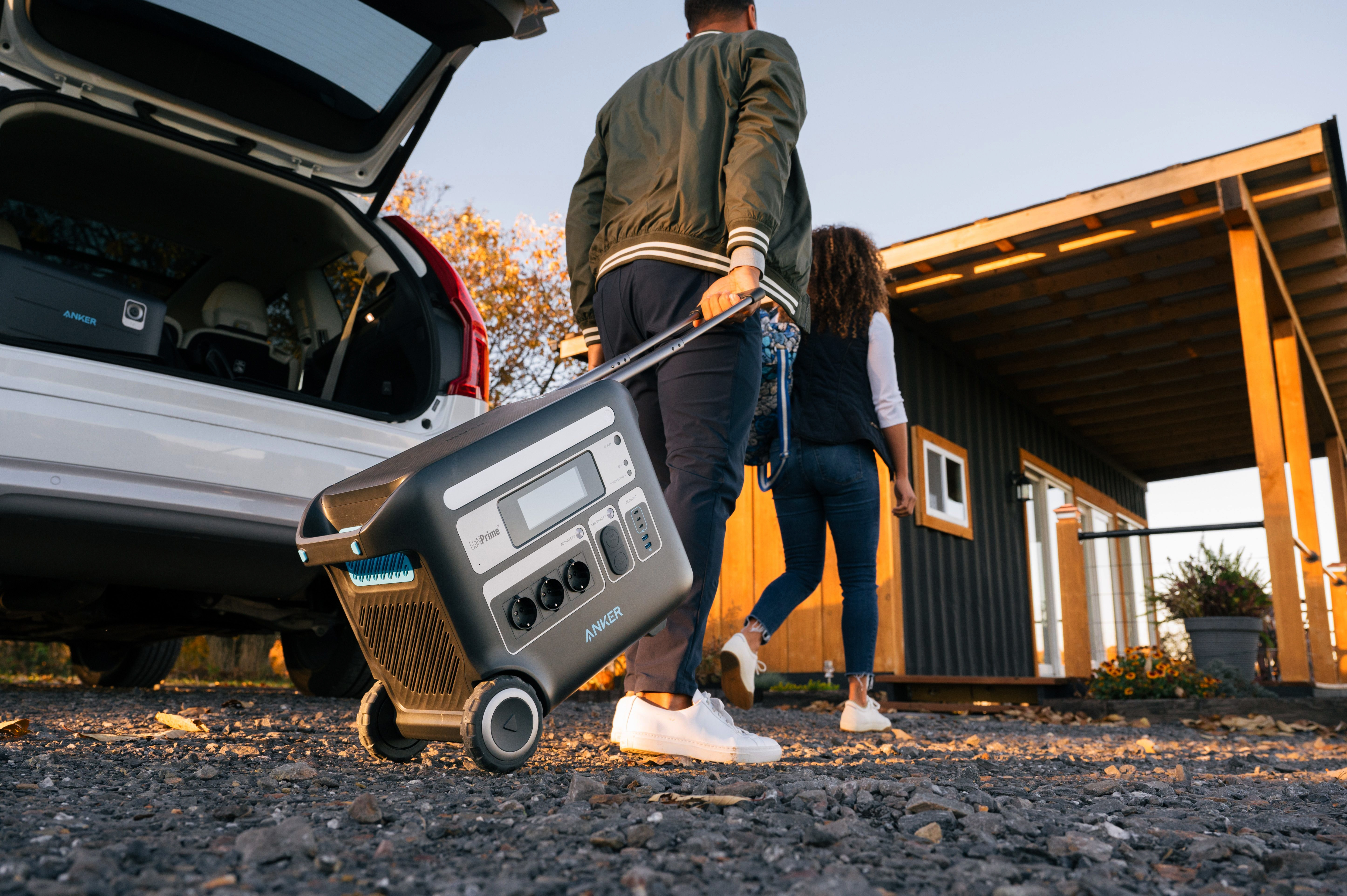 Dwie osoby idą w stronę domku, wyciągając z samochodu czarną elektrownię na kołach.