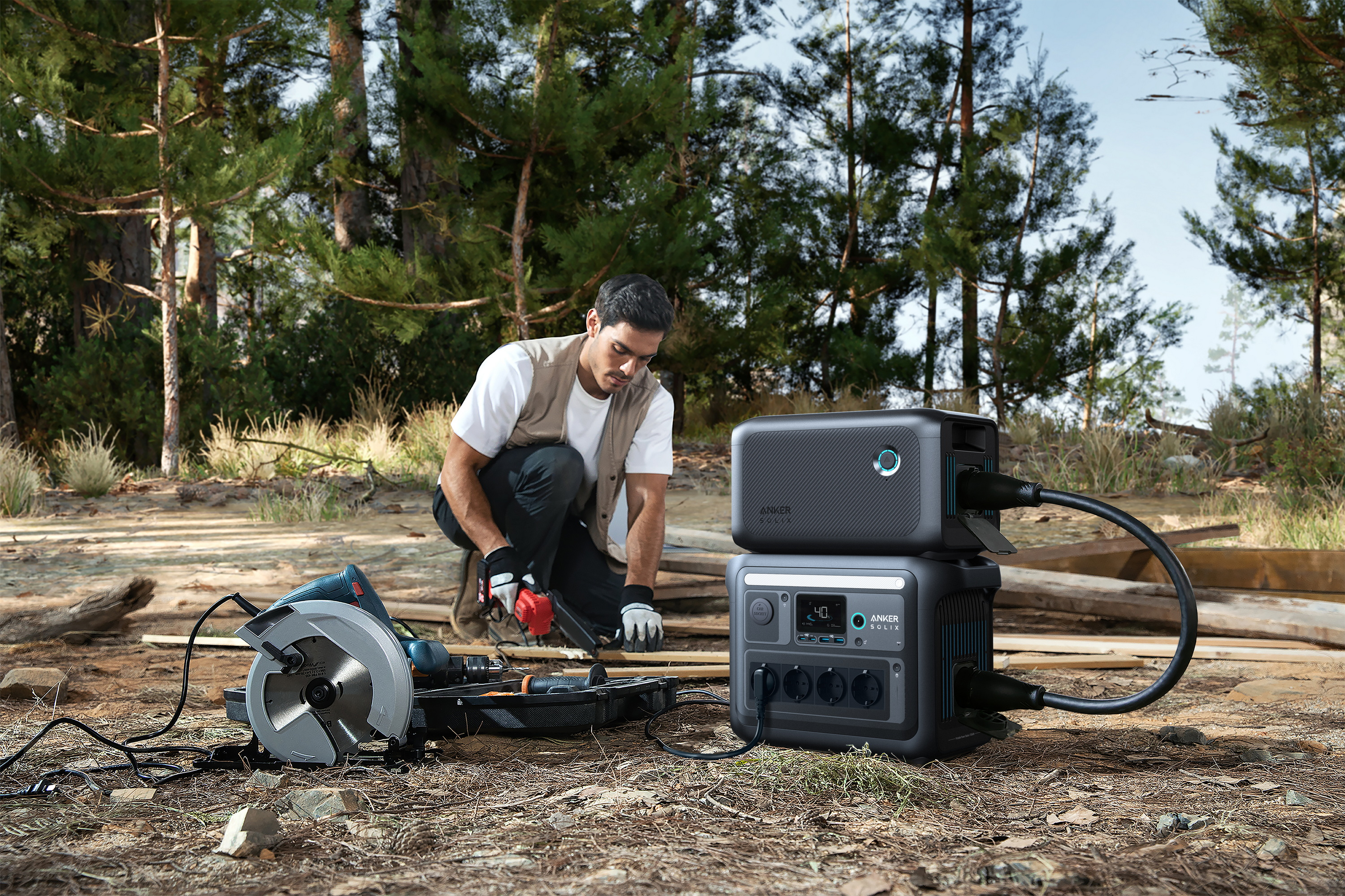 Mężczyzna kuca, pracuje nad drewnem piłą tarczową. Dwie stacje zasilania Anker w pobliżu. Las w tle.