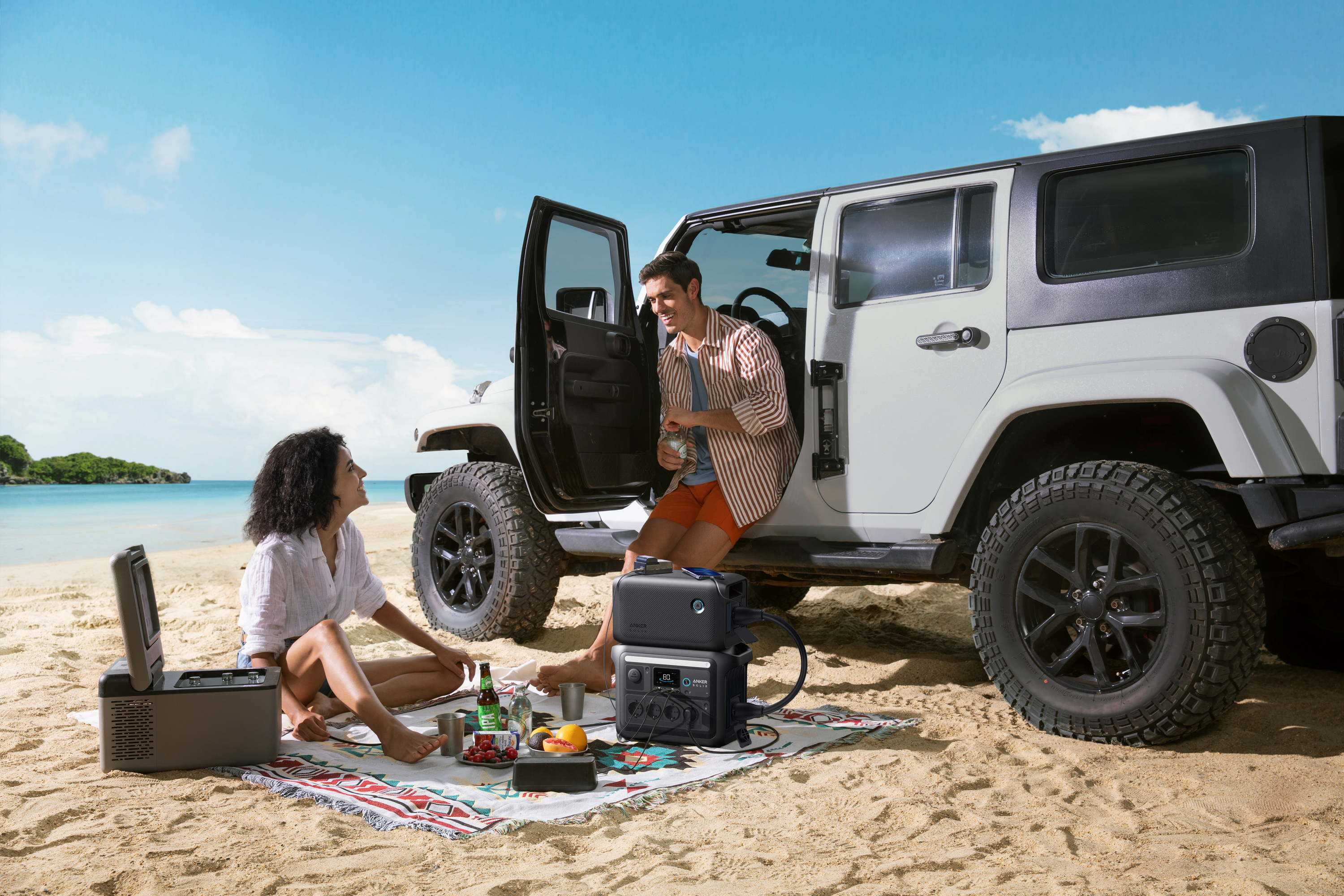 Dwie osoby na plaży z jeepem, lodówką i stacją zasilania.