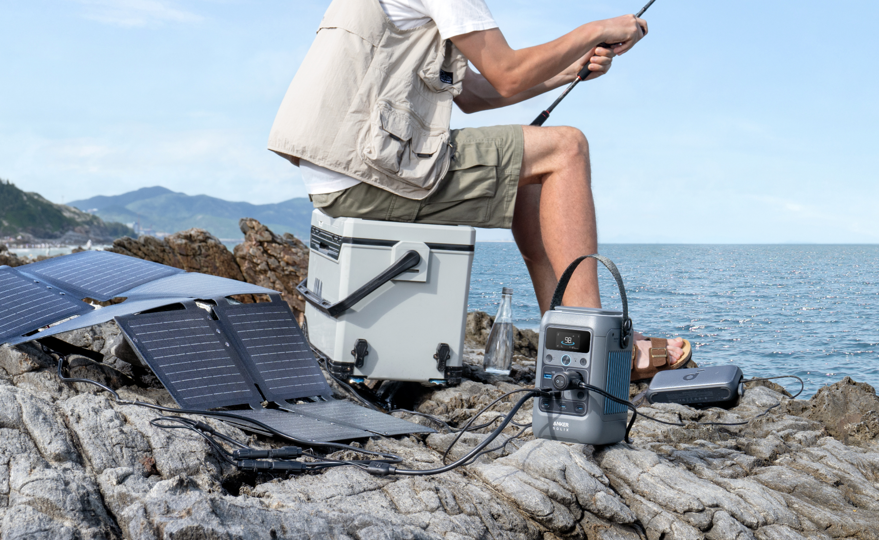 Osoba łowiąca ryby w pobliżu jeziora z panelem słonecznym, elektrownią i chłodziarką obok.