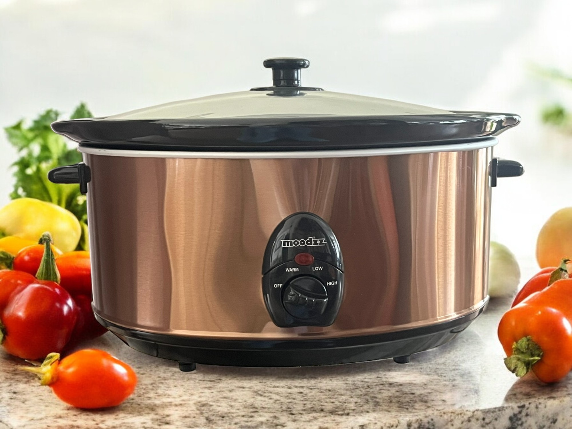 Een koperkleurige slowcooker met een zwart deksel staat op een aanrecht. Groenten staan op de voorgrond.