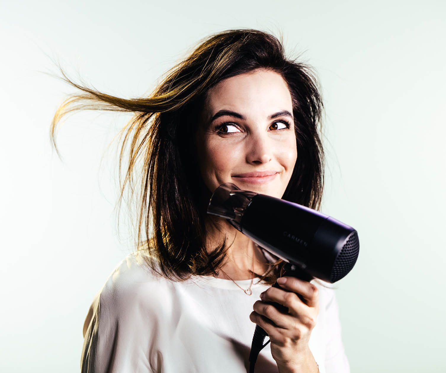 A woman holds a black hair dryer, looking off to the side with a smile, and her hair is blowing.