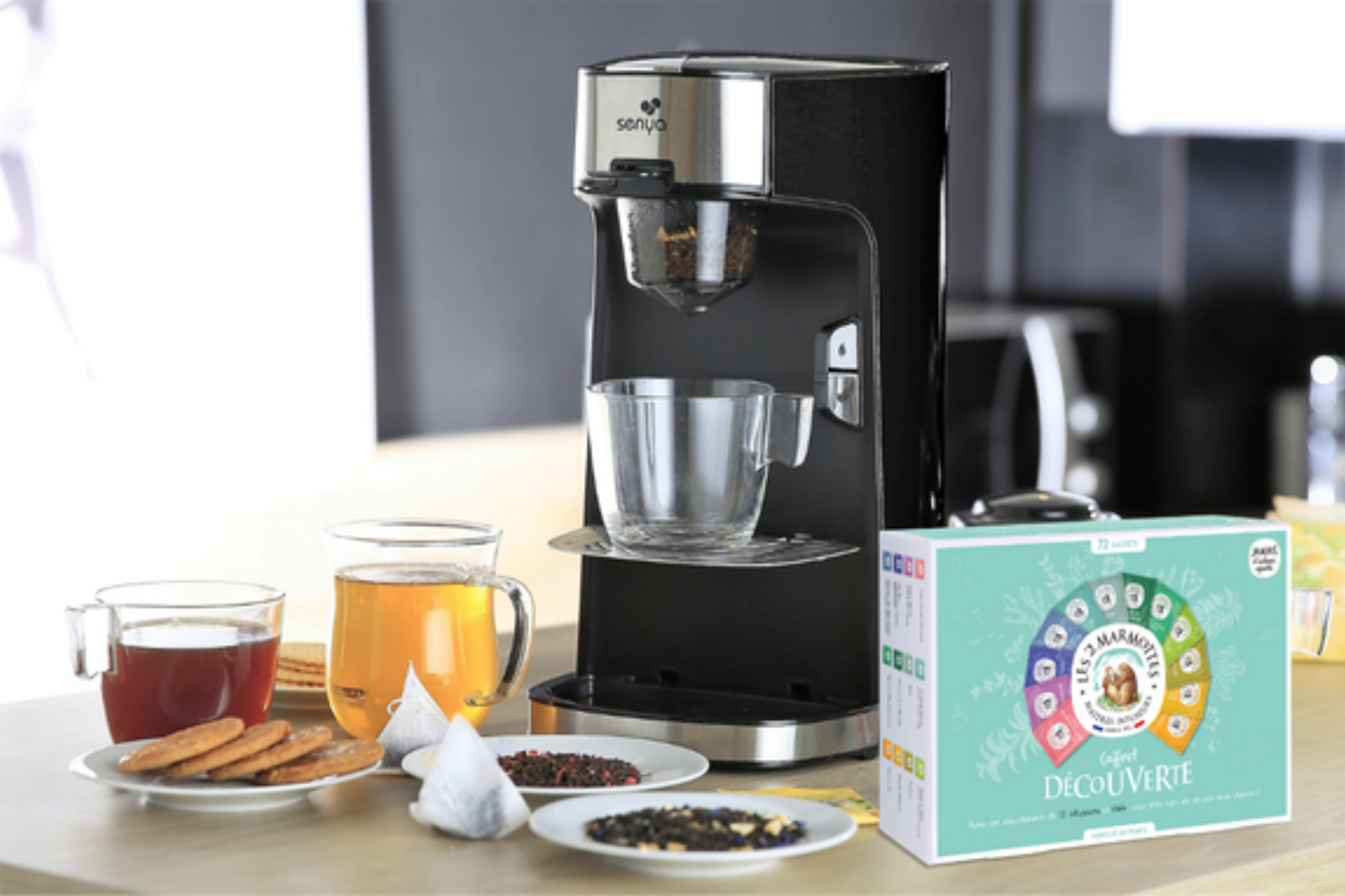 Macchina da caffè con tazza di vetro, tè, biscotti e una scatola di bustine di tè. Ambiente cucina.
