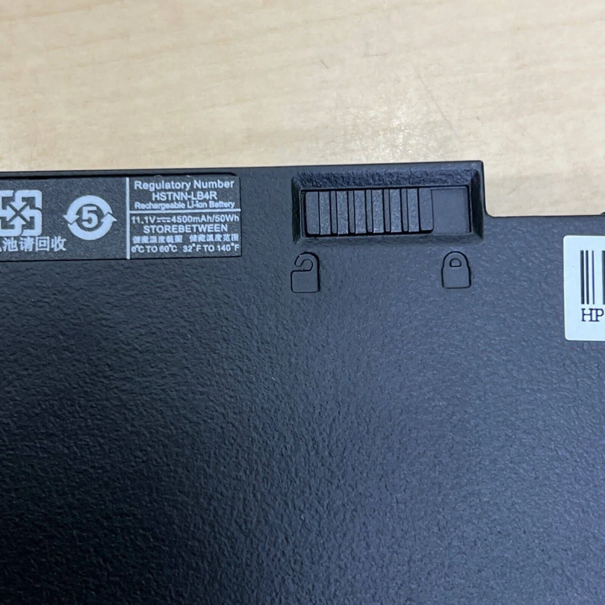 Czarny akumulator do laptopa z etykietą z tekstem i symbolami, na drewnianej powierzchni.