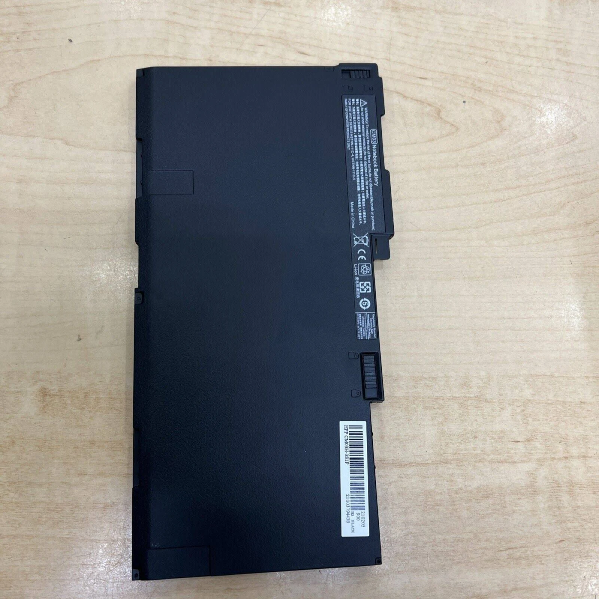 Czarna bateria do laptopa na drewnianej powierzchni. Widoczne etykiety i oznaczenia.