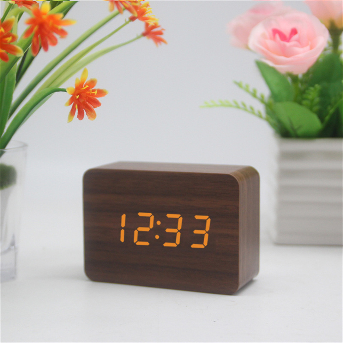 Holzblockuhr mit der Anzeige 12:33. Orangefarbene Ziffern. Hintergrund zeigt Blumen.